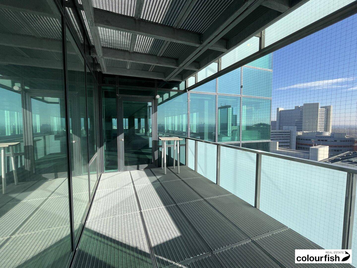 Geräumiger Balkon mit Metallboden und Glasgeländer, der einen Blick auf moderne Gebäude bietet.