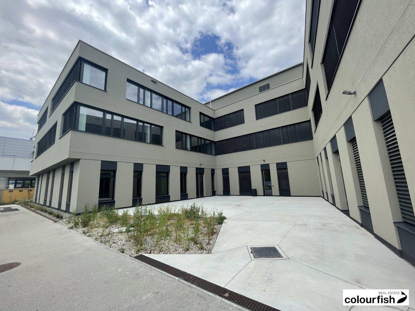 Ansicht eines modernen Gebäudekomplexes mit Innenhof und minimalistischer Bepflanzung unter bewölktem Himmel.