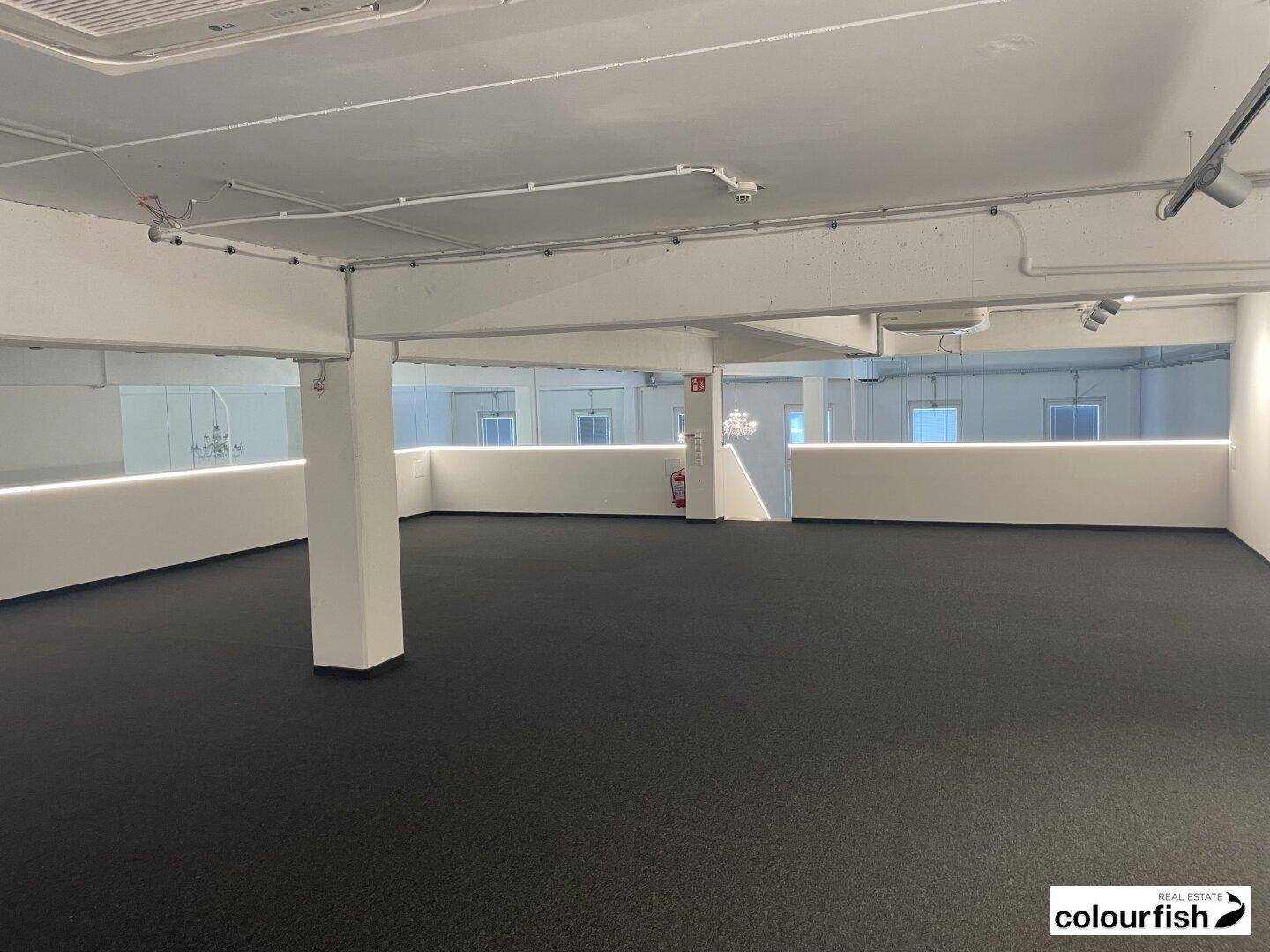 Großer, offener Bürobereich mit Säulen, dunklem Teppichboden und indirekter Beleuchtung entlang der Wände.