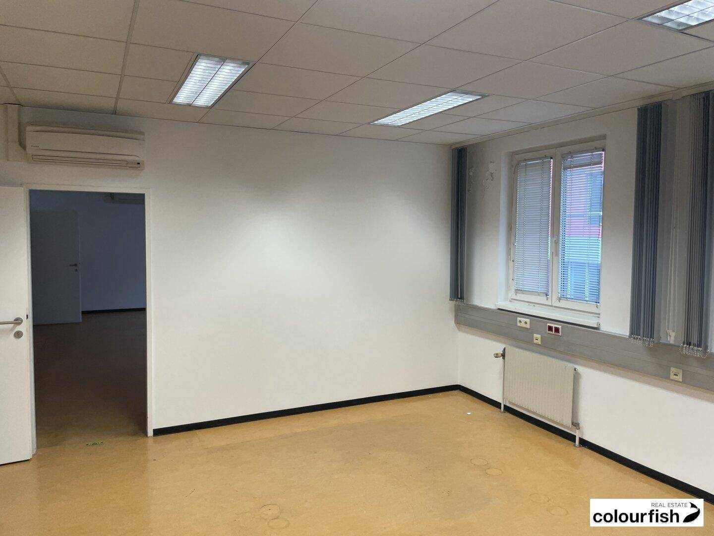 Leeres Bürozimmer mit Fenster, Heizkörper, abgehängter Decke und Klimaanlage, ideal für Einzelbüros.