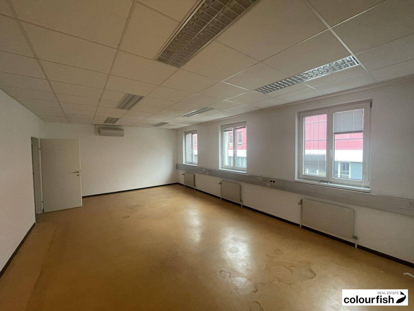 Geräumiges, leeres Büro mit mehreren Fenstern, Heizkörpern und Klimaanlage, bietet viel Tageslicht.