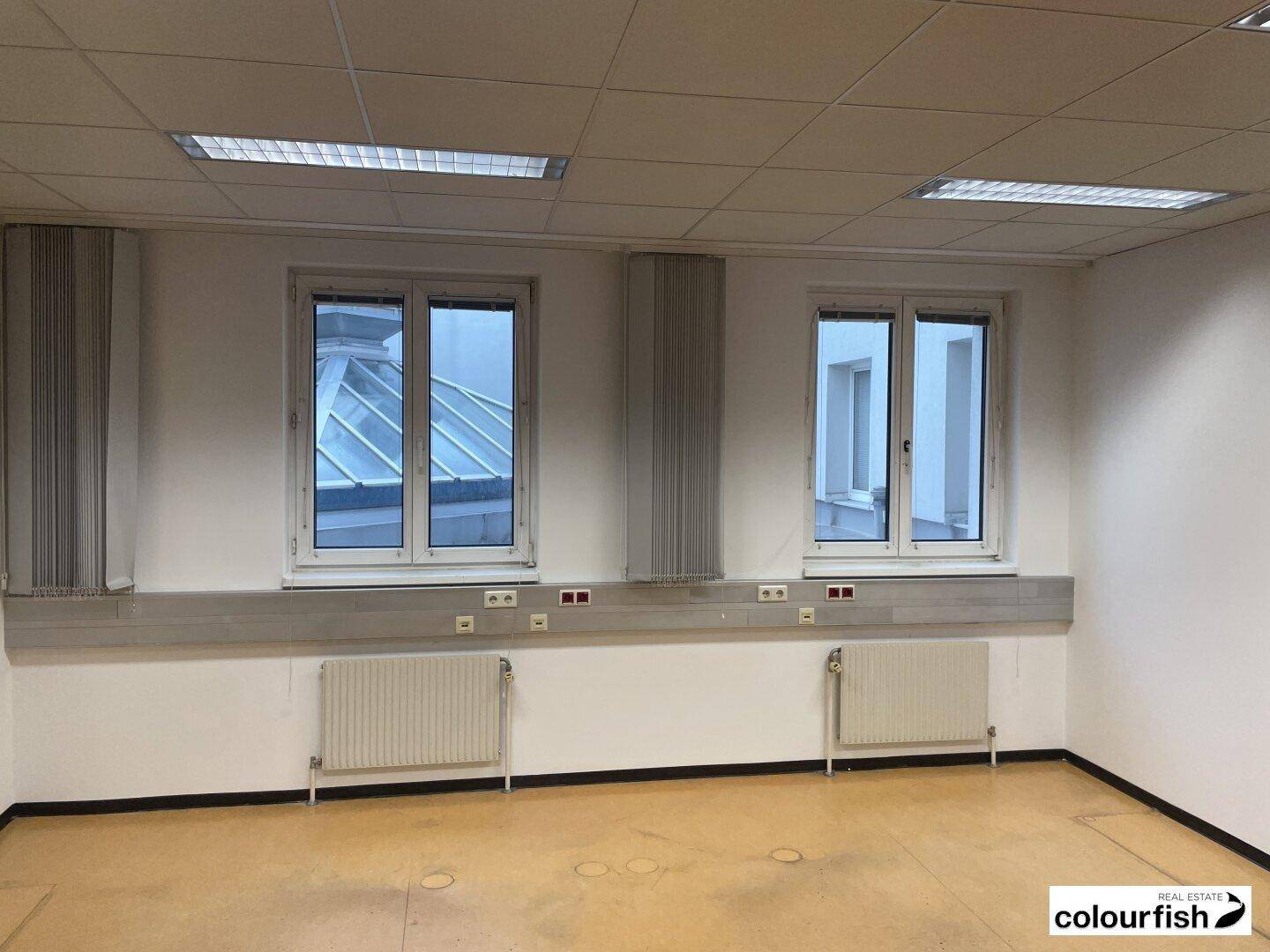 Helles Bürozimmer mit zwei Fenstern und Blick auf ein Dach, ausgestattet mit Heizkörpern und Rasterdecke.