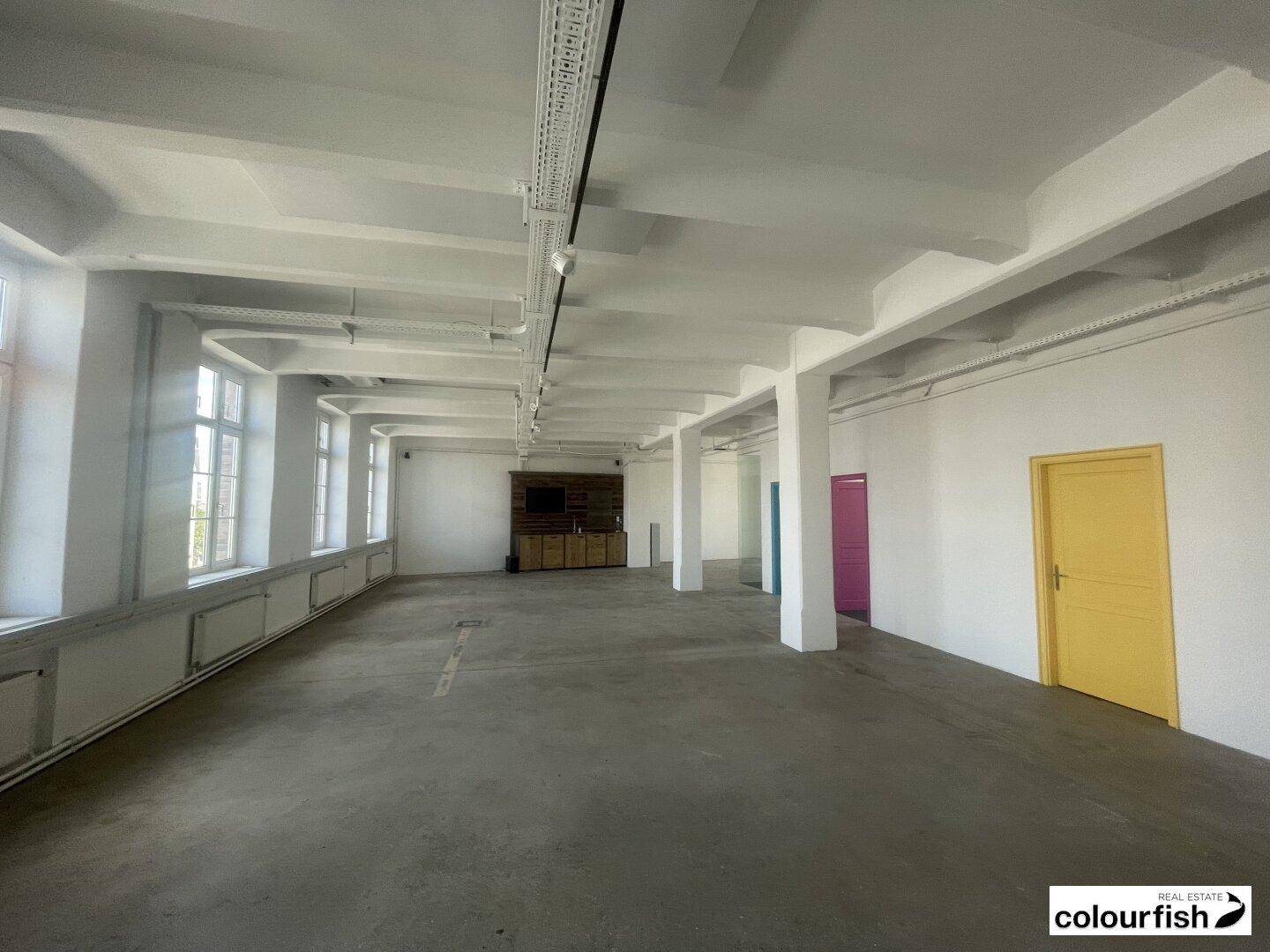 Weitläufiger Büroraum mit Betonboden, hohen Decken und farbigen Türen entlang der weißen Wand.
