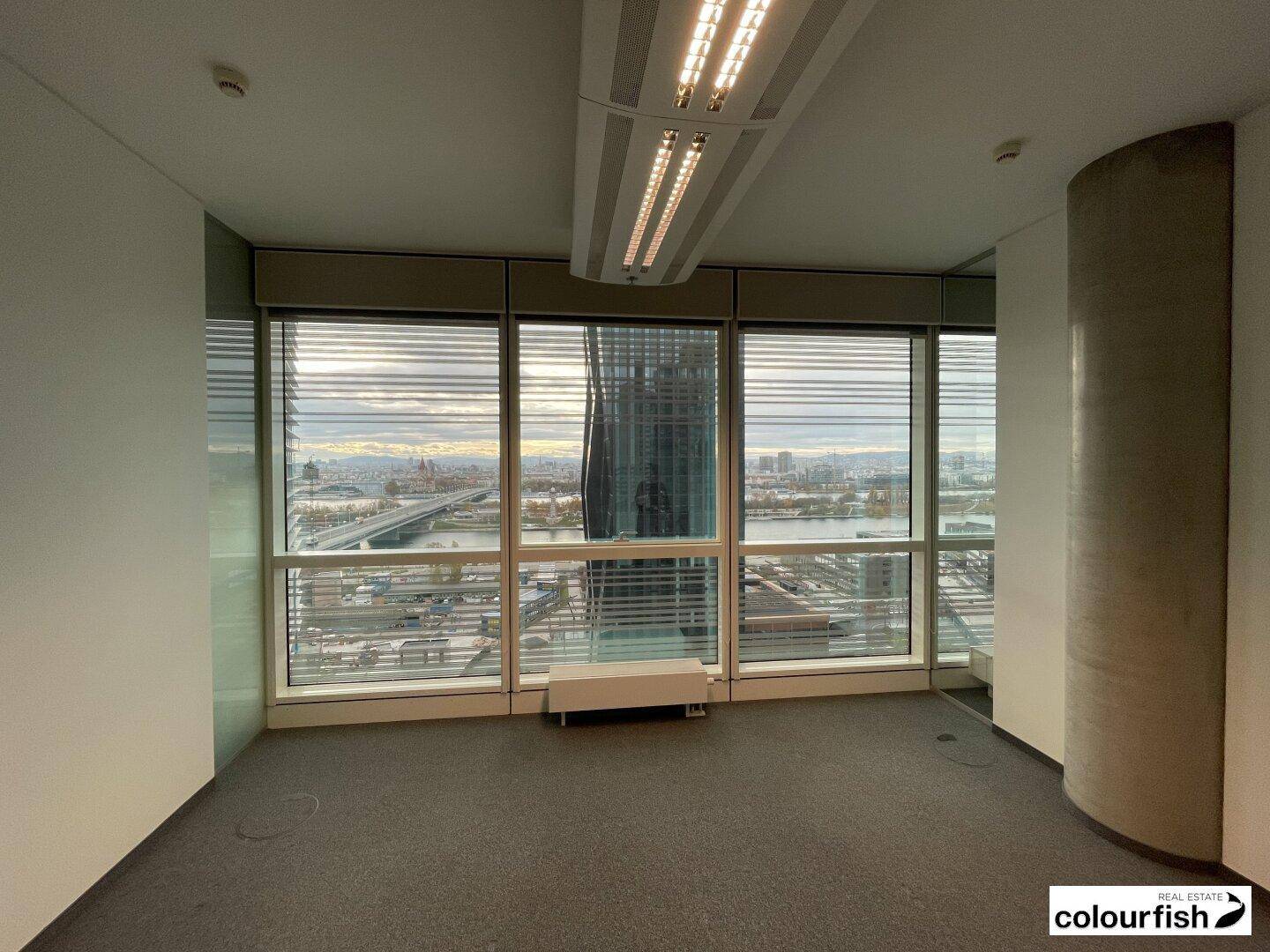Helles Büro mit Teppichboden und breiten Fenstern, die einen Panoramablick auf die städtische Umgebung ermöglichen.