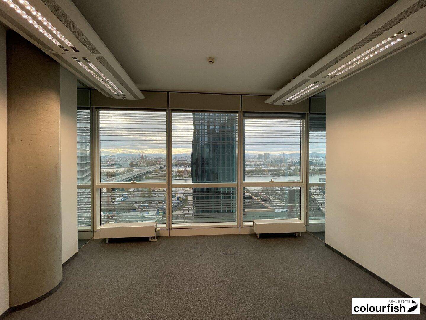 Leeres Büro mit Teppichboden und großen Fenstern, die eine weite Aussicht auf die Stadtlandschaft bieten.