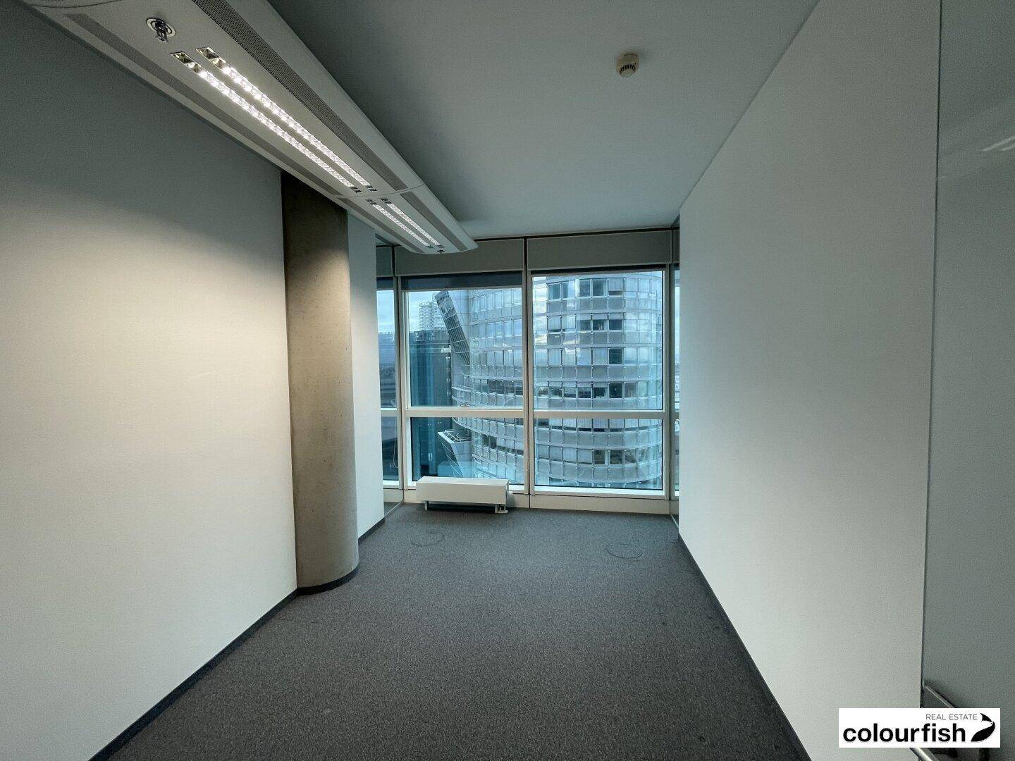 Kleineres Büro mit Teppichboden und großen Fenstern, die einen Blick auf benachbarte Gebäude ermöglichen.