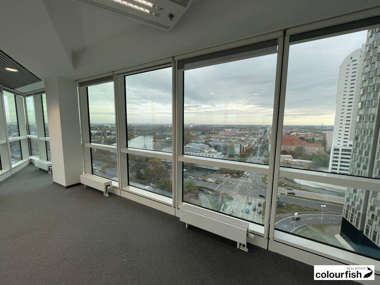 Bürofläche mit Teppichboden und großen Fenstern, die einen weiten Blick auf die Stadt und den Fluss ermöglichen.
