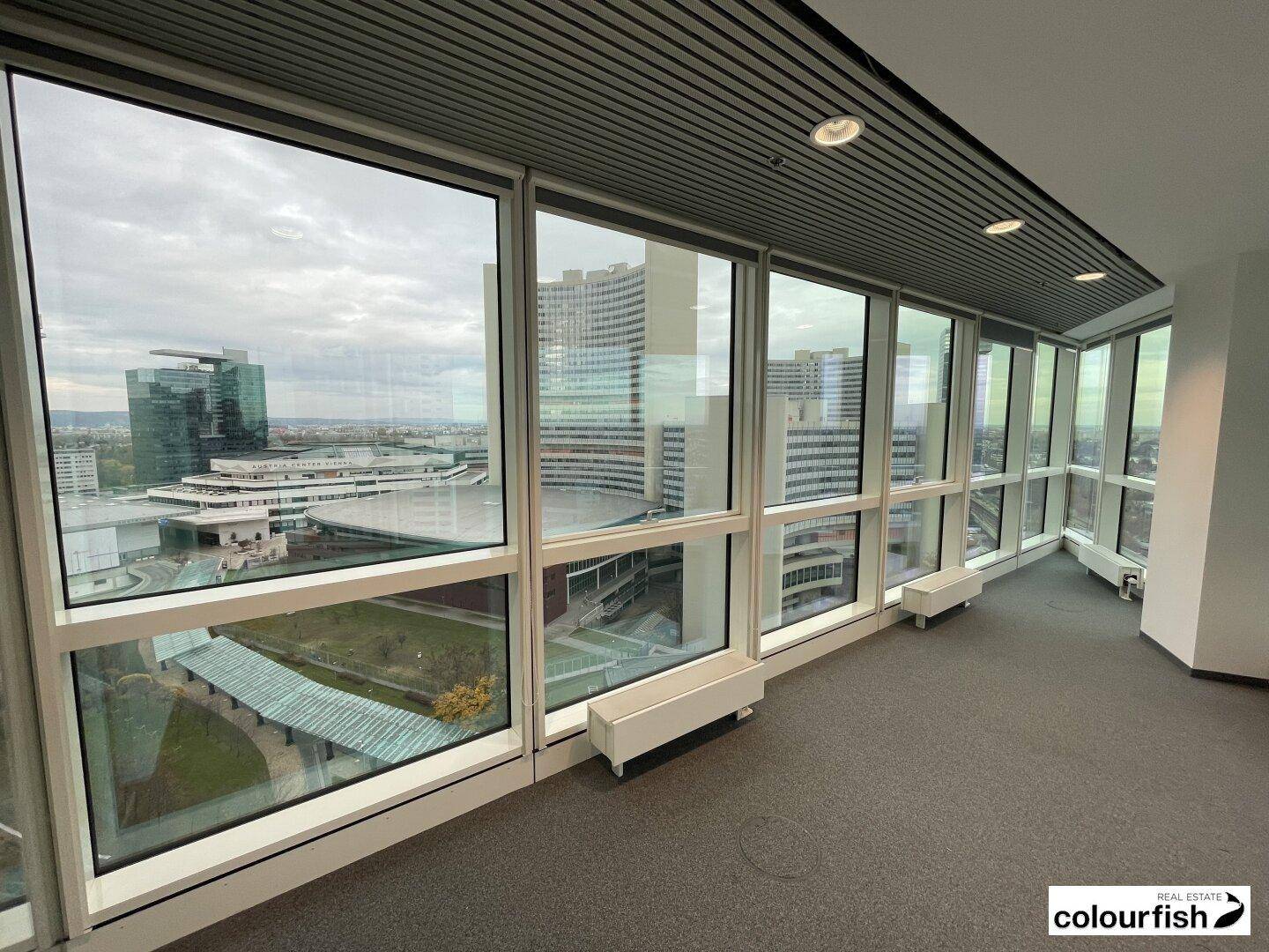 Büro mit Teppichboden und raumhohen Fenstern, die einen weiten Blick auf die umliegenden Gebäude bieten.
