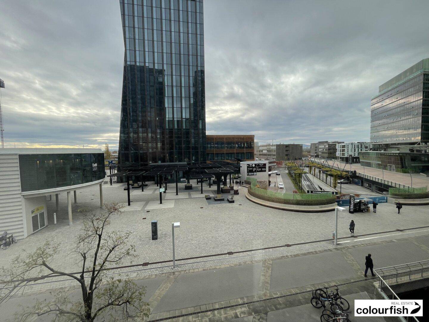 Blick von oben auf einen modernen Stadtplatz mit umliegenden Hochhäusern und städtischem Treiben.