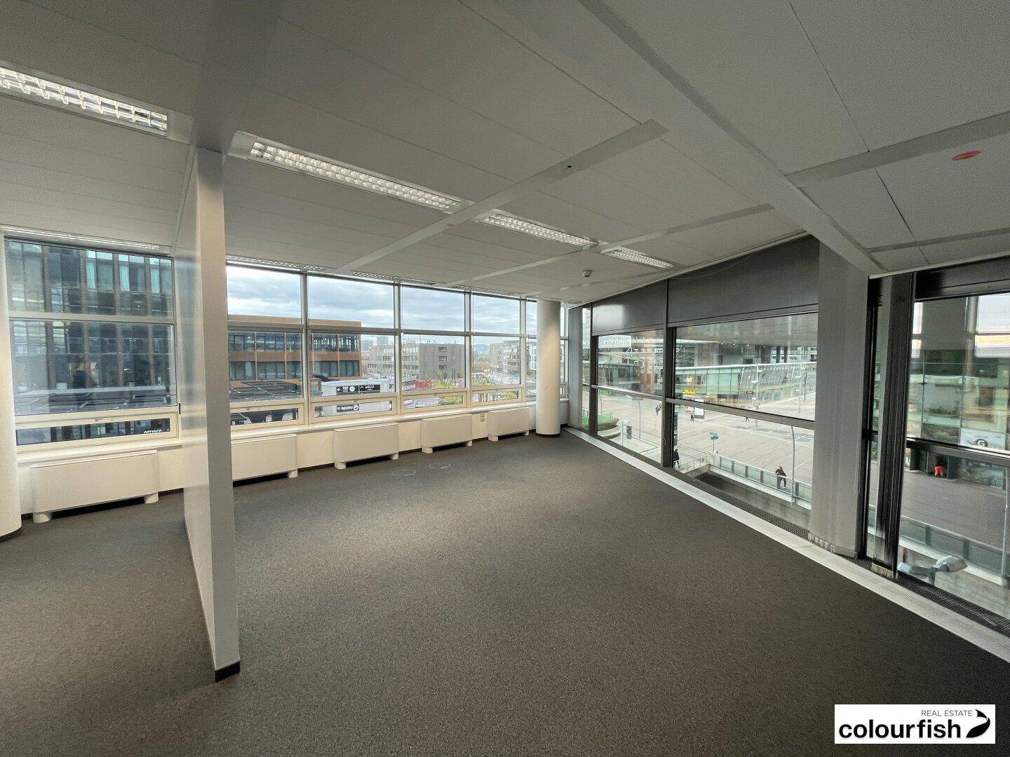 Helles Eckbüro mit Teppichboden und Panoramafenstern, die einen weiten Blick über die Stadt bieten.