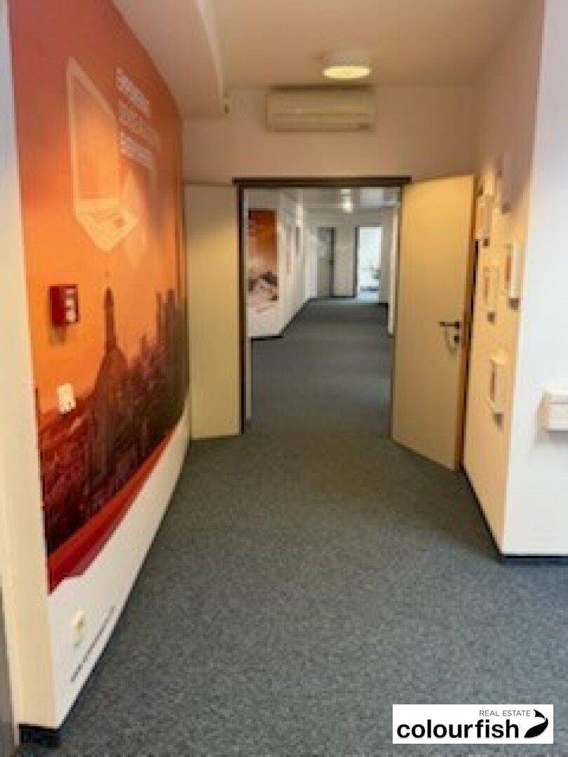 Büroflur mit Teppichboden, einer Wanddekoration und einer Klimaanlage über dem Türrahmen.