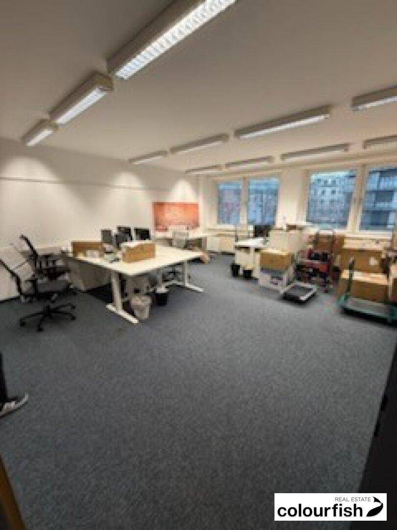 Großzügiger Büroraum mit mehreren Arbeitsplätzen, Teppichboden und Fenstern für Tageslicht.