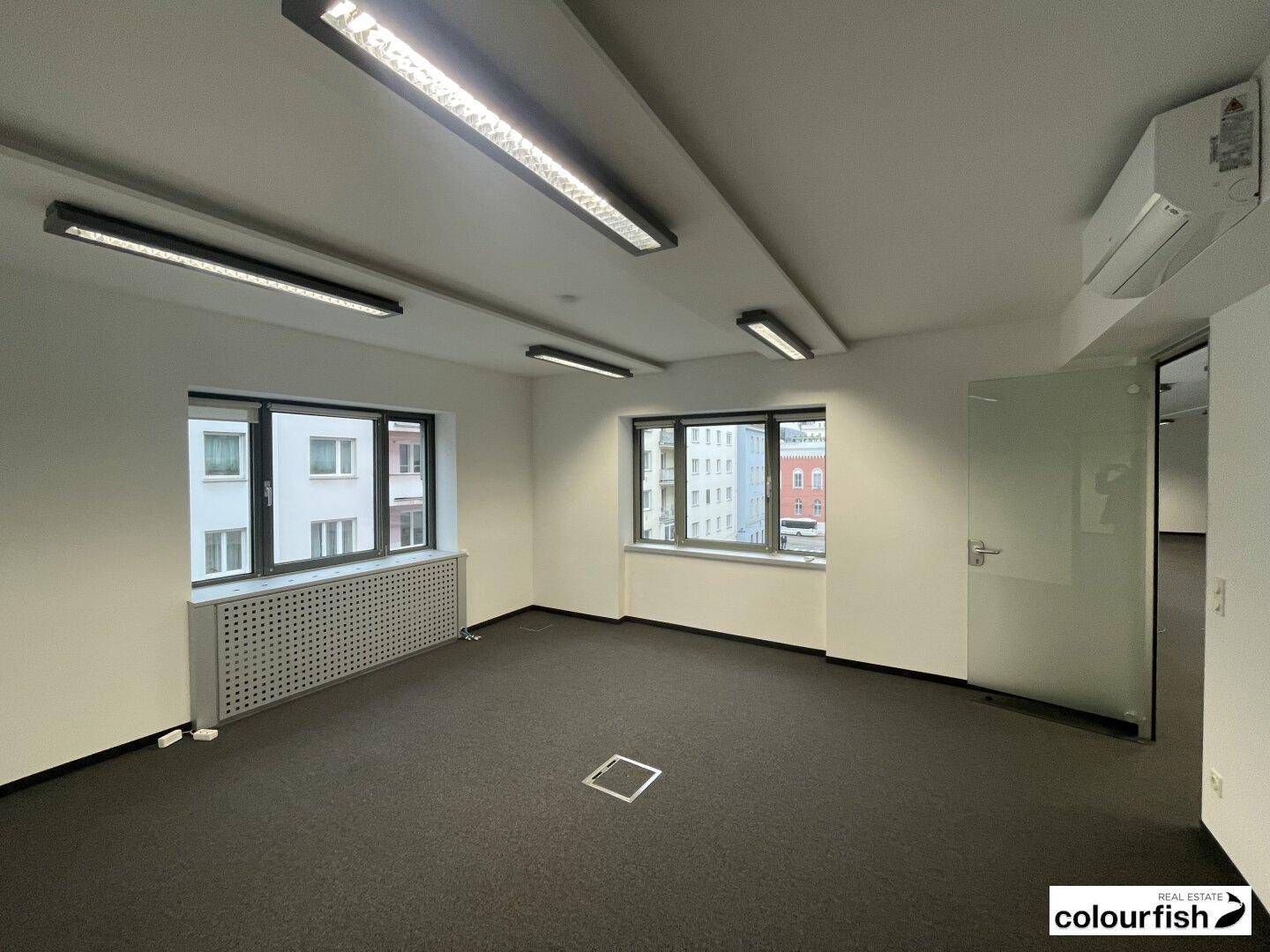 Heller Büroraum mit Teppichboden, großen Fenstern und einer Glastür zu einem weiteren Bereich.