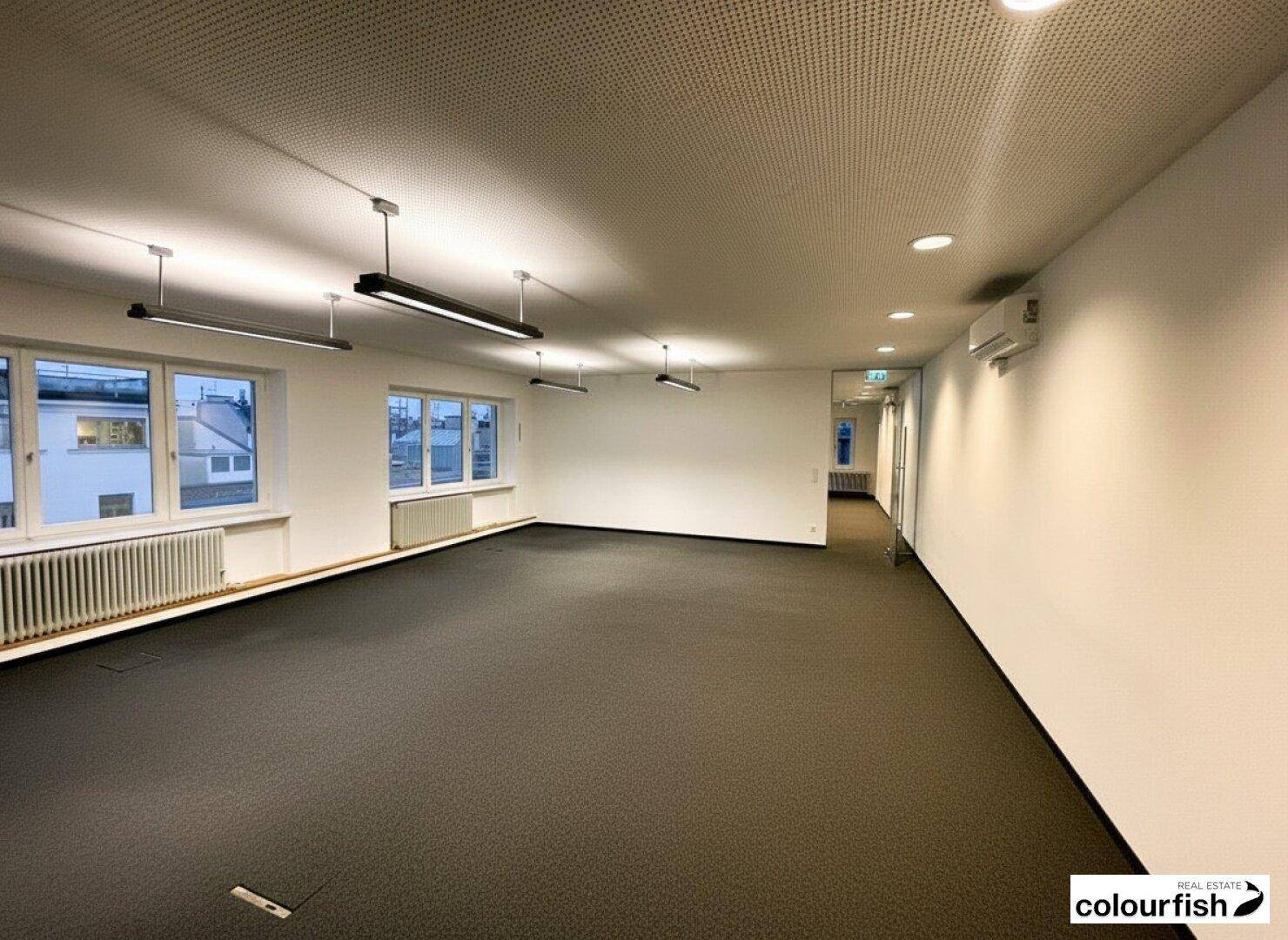 Großer, leerer Büroraum mit Teppichboden, vielen Fenstern und moderner Deckenbeleuchtung sowie Klimaanlage.