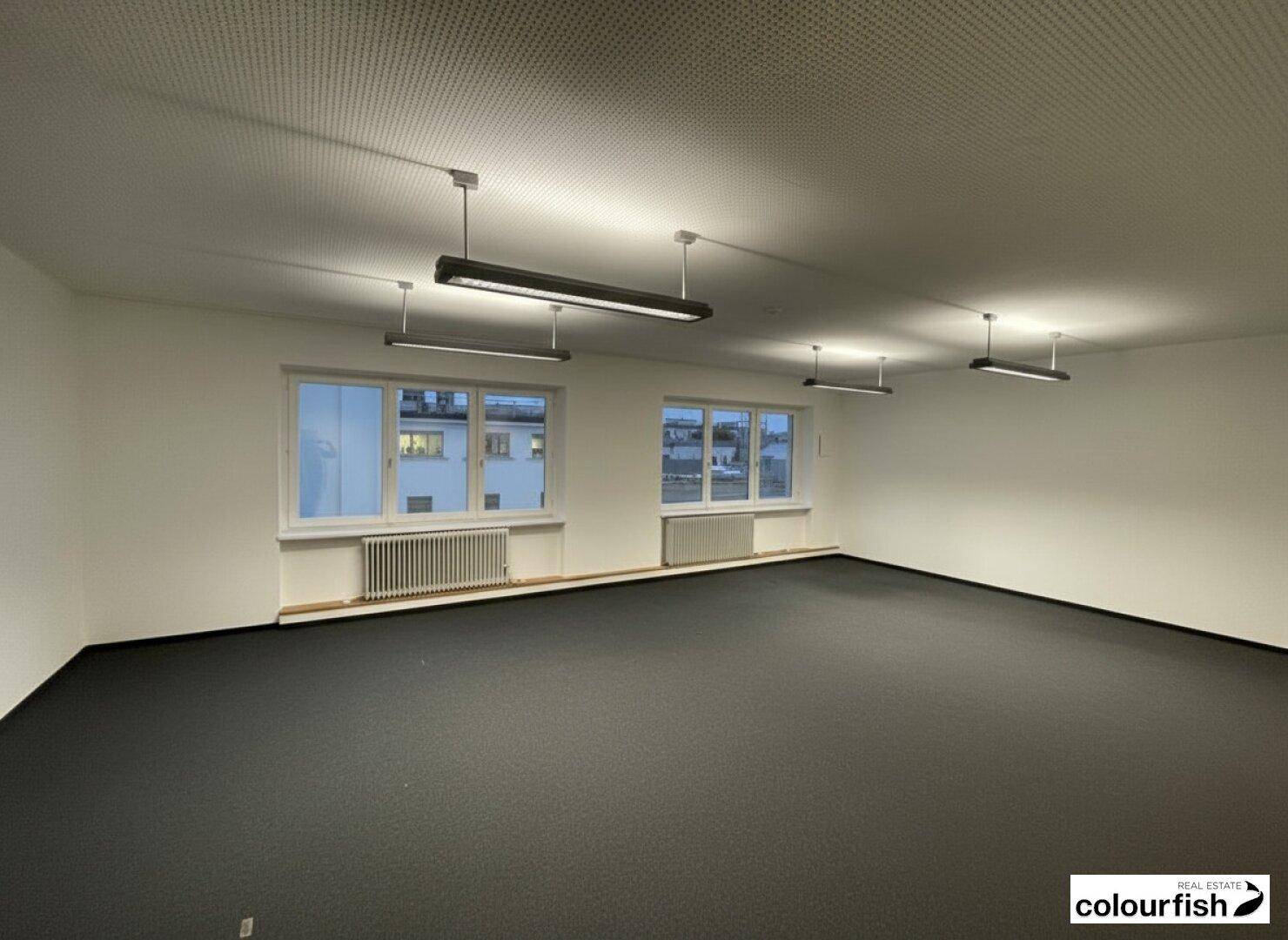 Geräumiger Bürobereich mit dunklem Teppichboden, großen Fenstern und moderner Beleuchtung an der Decke.