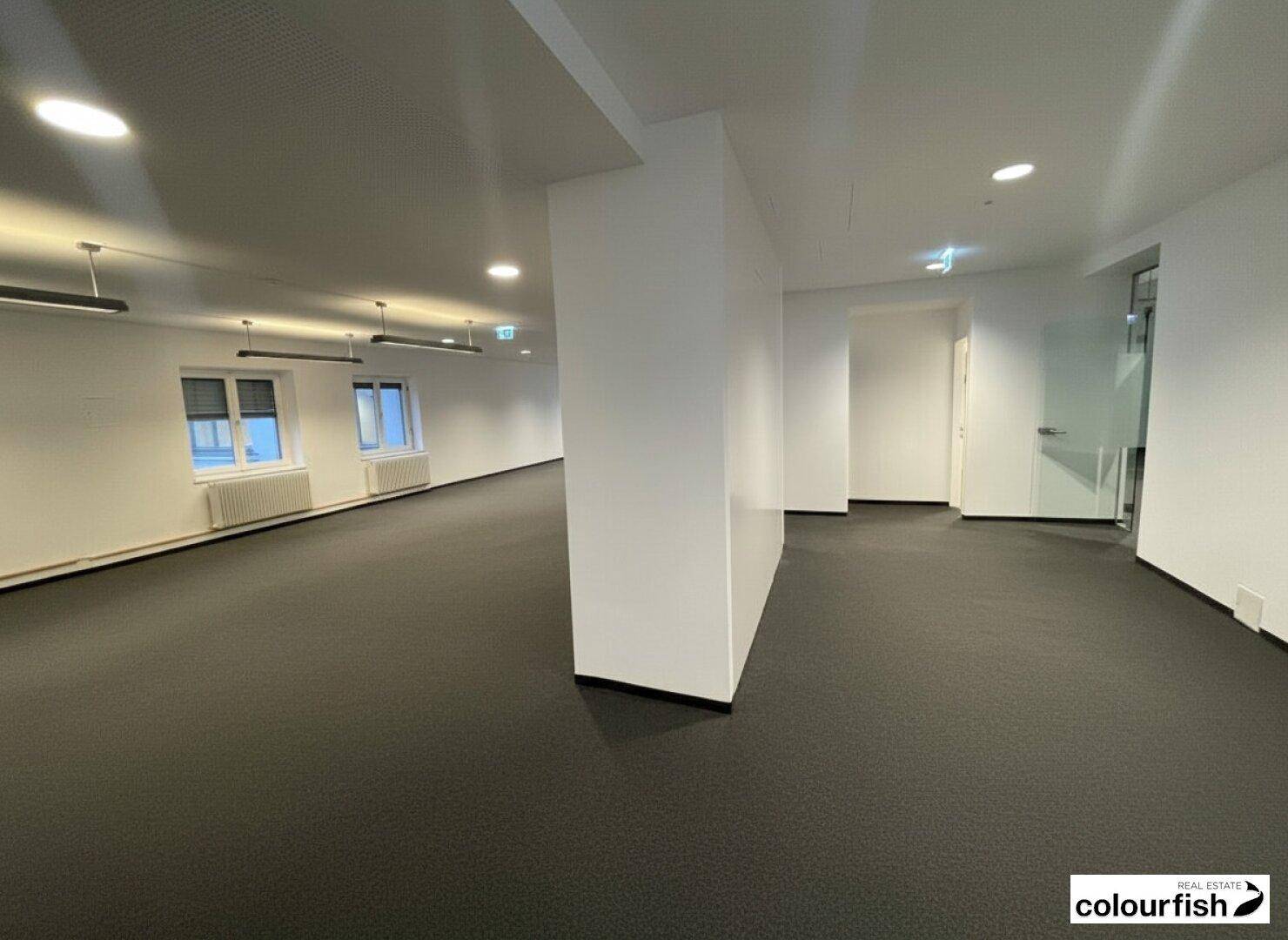 Offener Bürobereich mit Säule, Teppichboden, Deckenbeleuchtung und Blick auf weitere Räume.