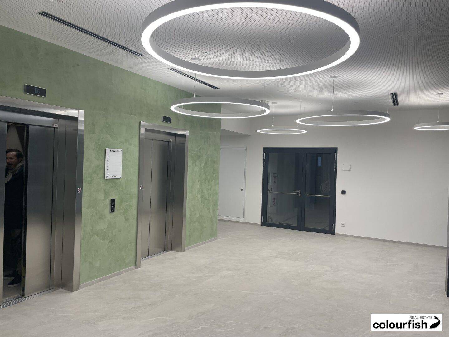 Moderne Lobby mit zwei Aufzügen, grüner Akzentwand, hellgrauem Fliesenboden und runden LED-Deckenleuchten.