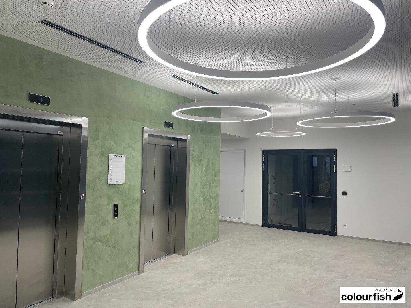 Helle und moderne Empfangshalle mit Edelstahlaufzügen, strukturierter grüner Wand und stilvollen Deckenleuchten.