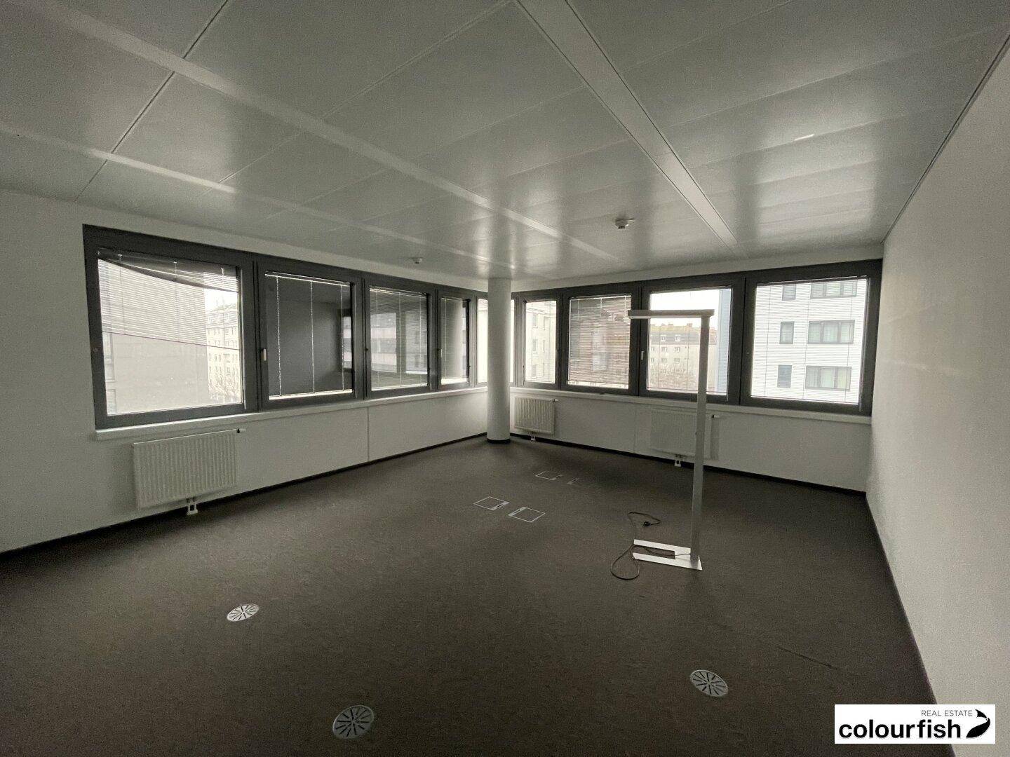 Heller Eckbüroraum mit Teppichboden, großen Fenstern an zwei Seiten und Blick auf die städtische Umgebung.