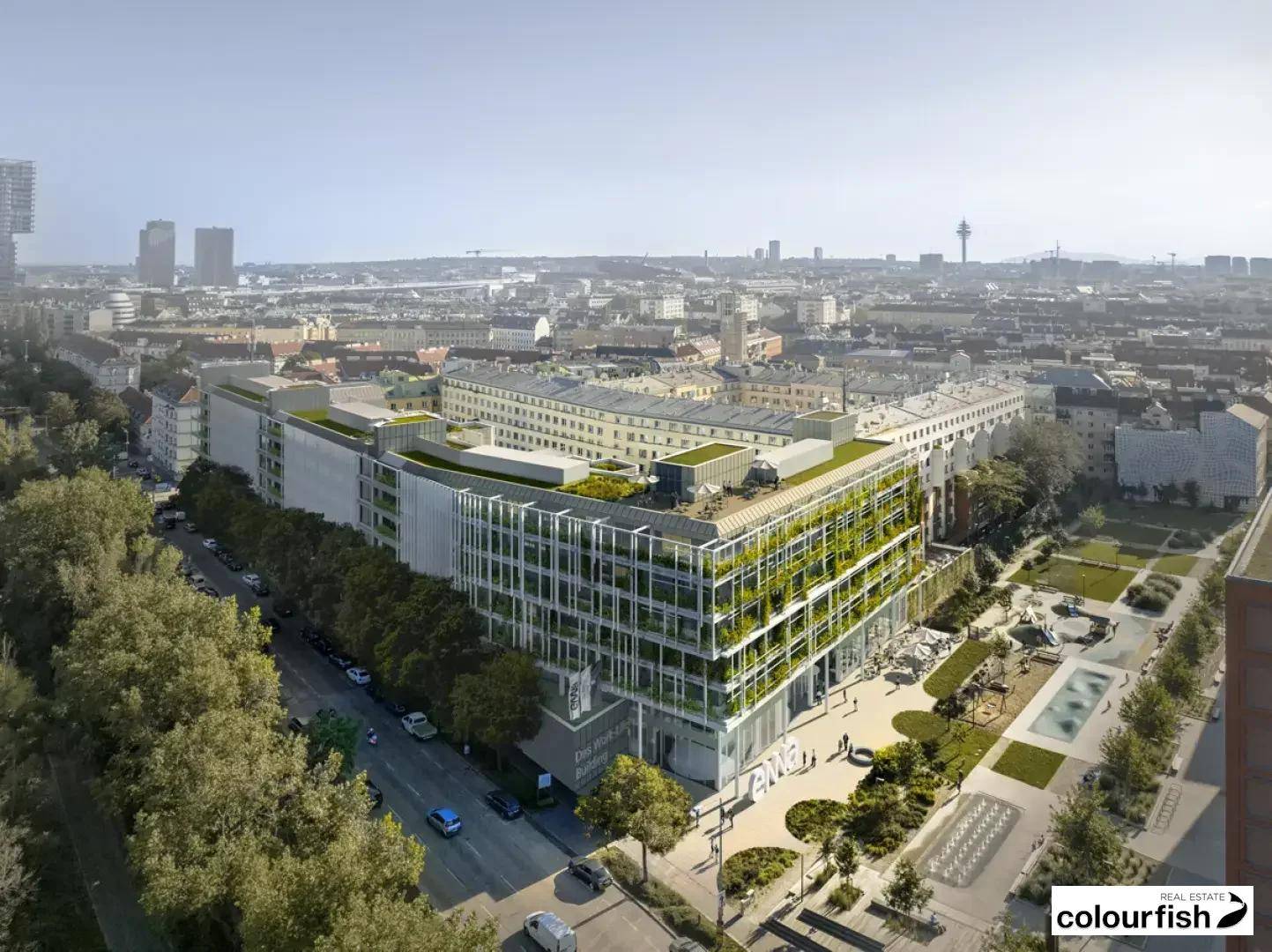 Luftaufnahme eines modernen Gebäudes mit begrünten Fassaden und Dachflächen, umgeben von einer städtischen Landschaft und einem Park.