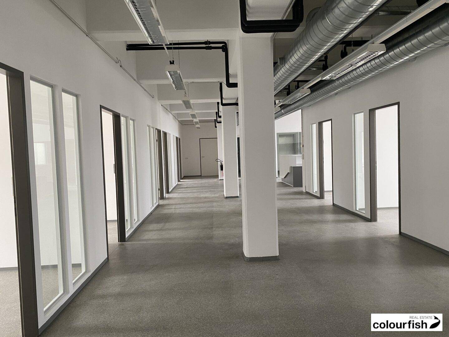 Langer, heller Bürogang mit vielen Türen zu einzelnen Büros und sichtbaren Lüftungsrohren an der Decke, modern gestaltet.