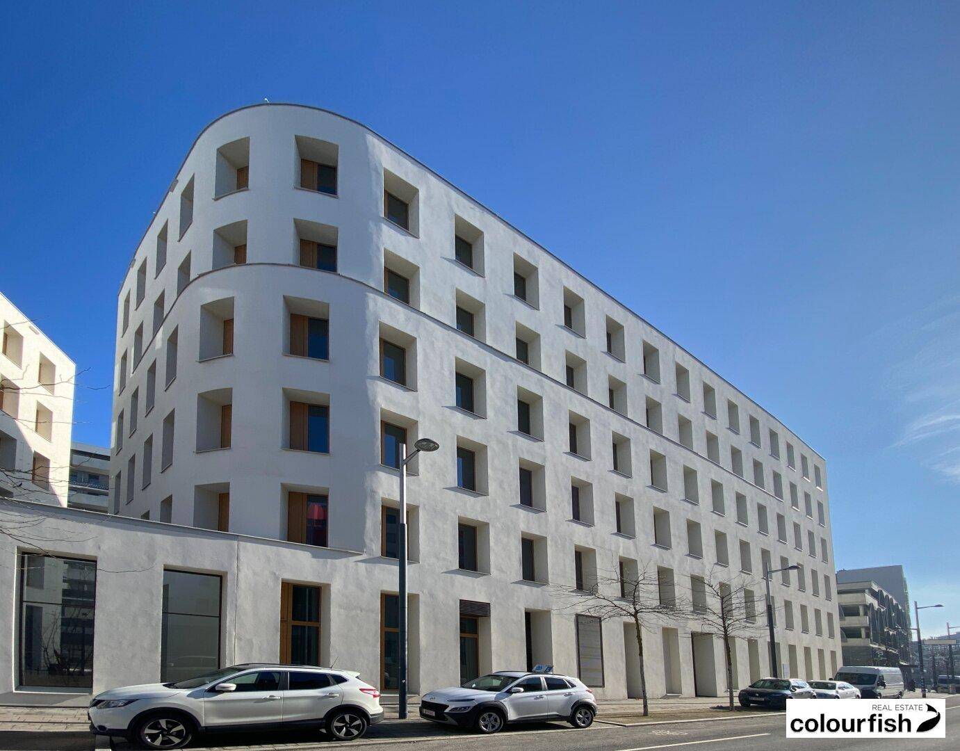 Weitläufiges, modernes Gebäude mit weißer Fassade und vielen Fenstern an einer städtischen Straße.
