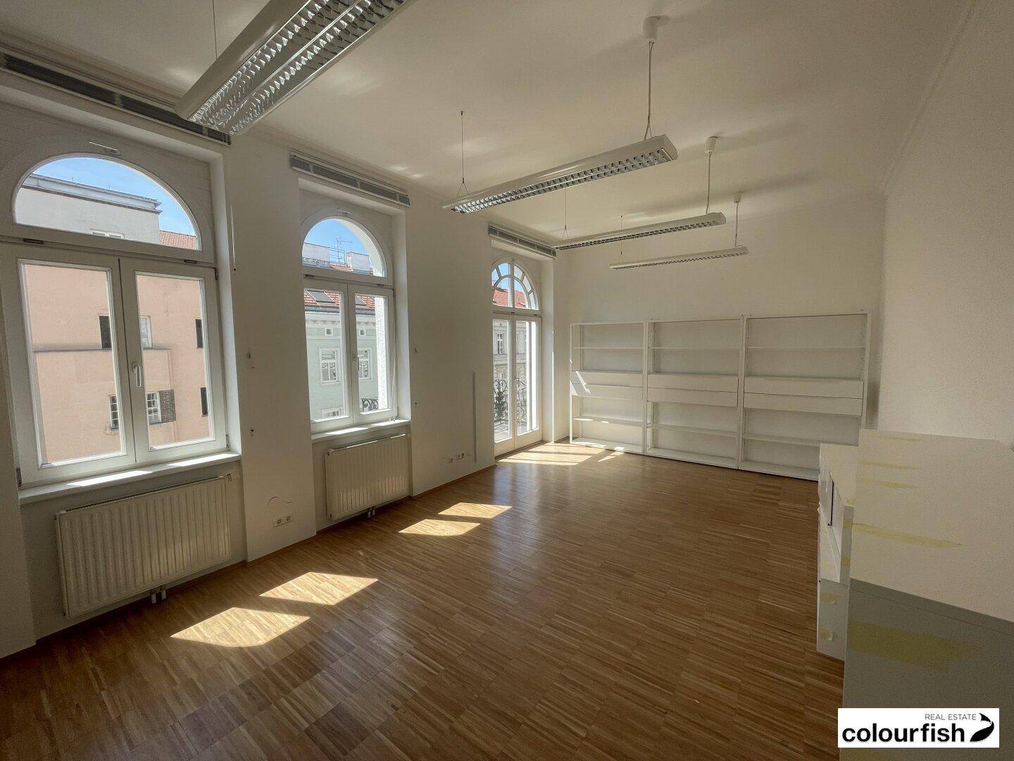 Heller Büroraum mit Holzboden, großen Bogenfenstern und integrierten Regalen, ideal für gewerbliche Nutzung.