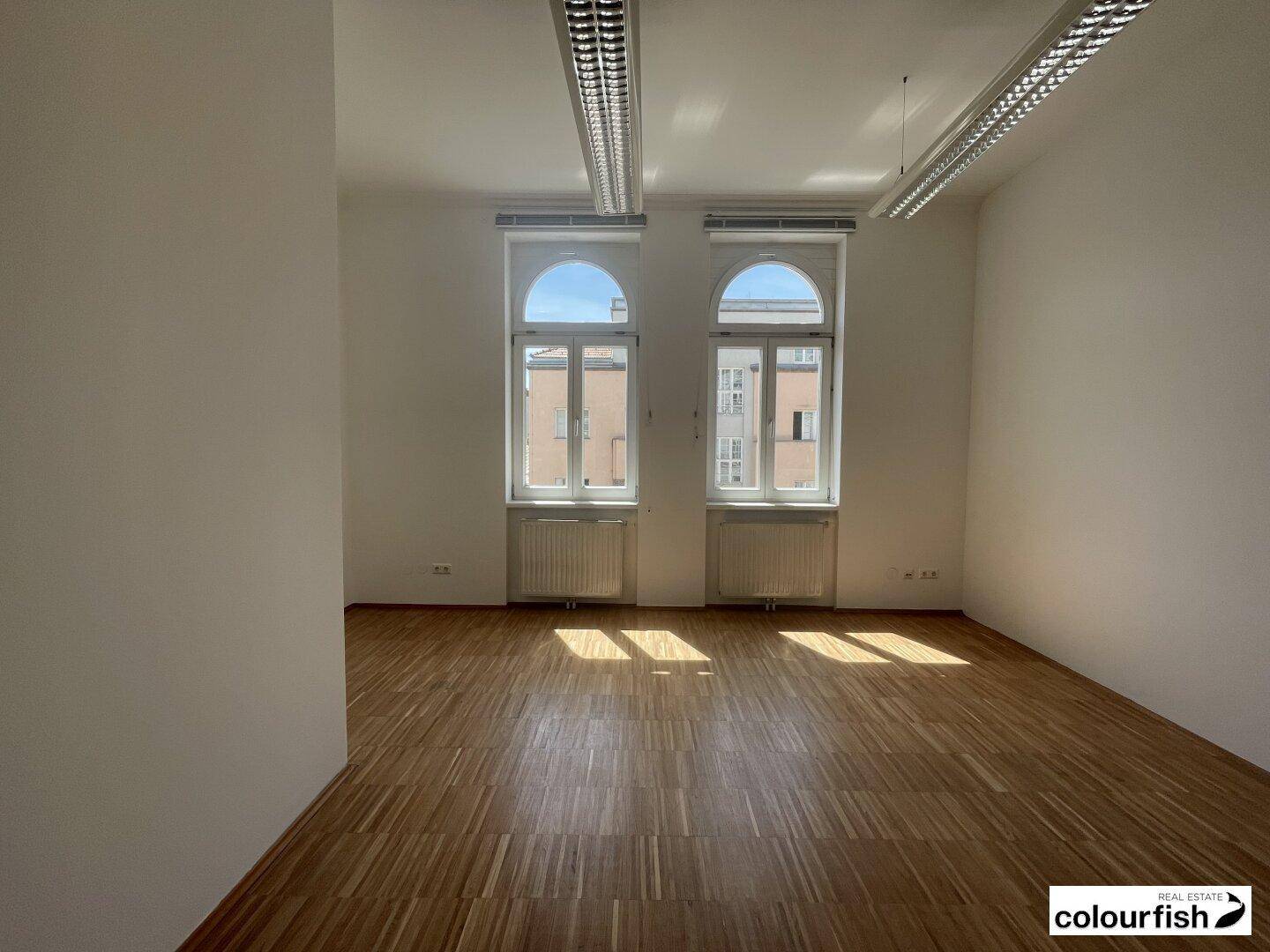 Geräumiger Büroraum mit zwei großen Bogenfenstern, Holzboden und viel Tageslicht, bereit zur Einrichtung.