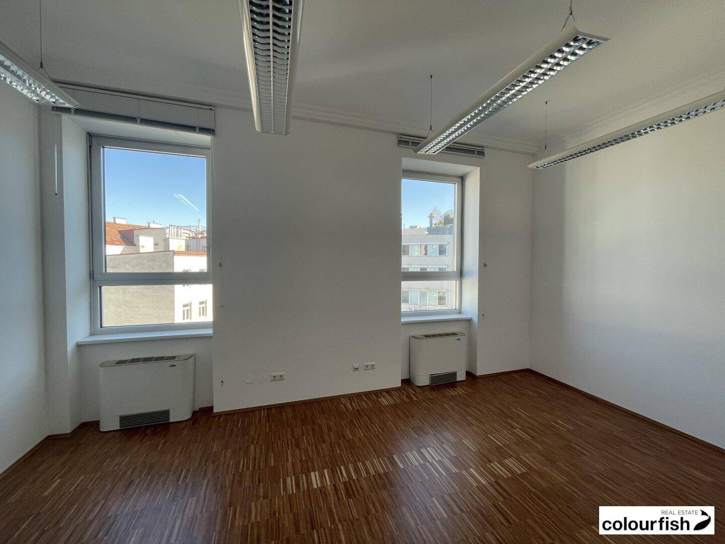 Heller Büroraum mit zwei großen Fenstern, Holzboden und effizienter Deckenbeleuchtung, ideal für Arbeitsplätze.
