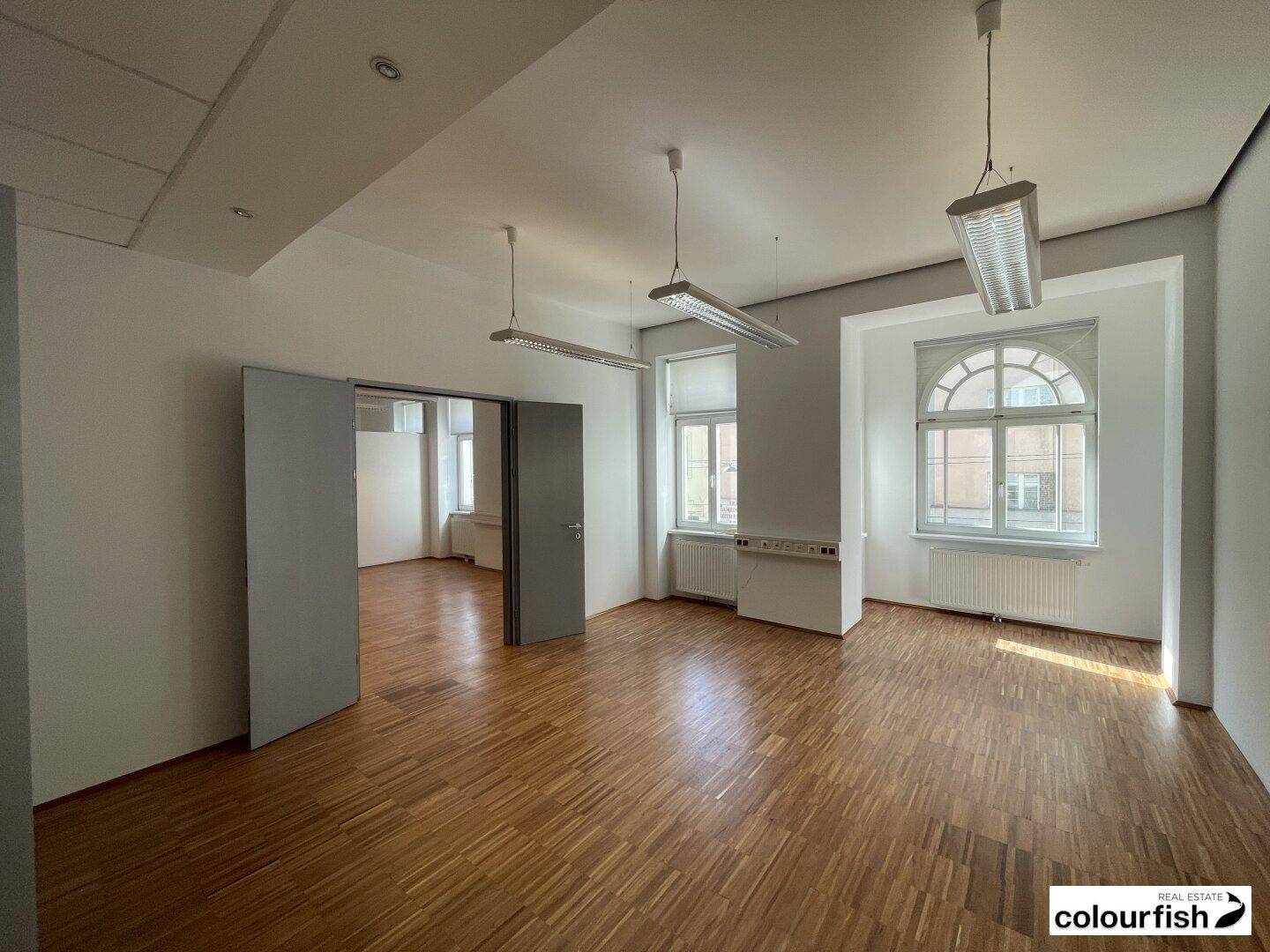 Geräumiger Büroraum mit elegantem Holzparkett, hohen Fenstern und Zugang zu weiteren Räumen.