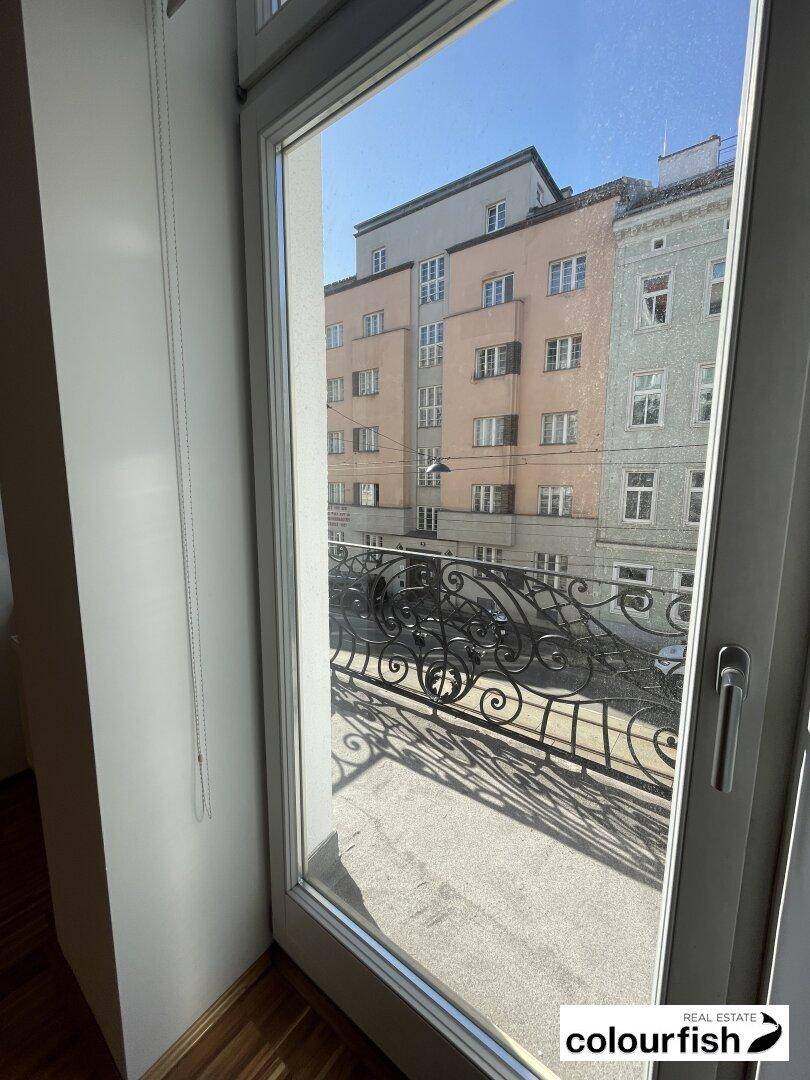 Blick vom Innenraum auf einen kleinen Balkon mit schmiedeeisernem Geländer und Stadtansicht.