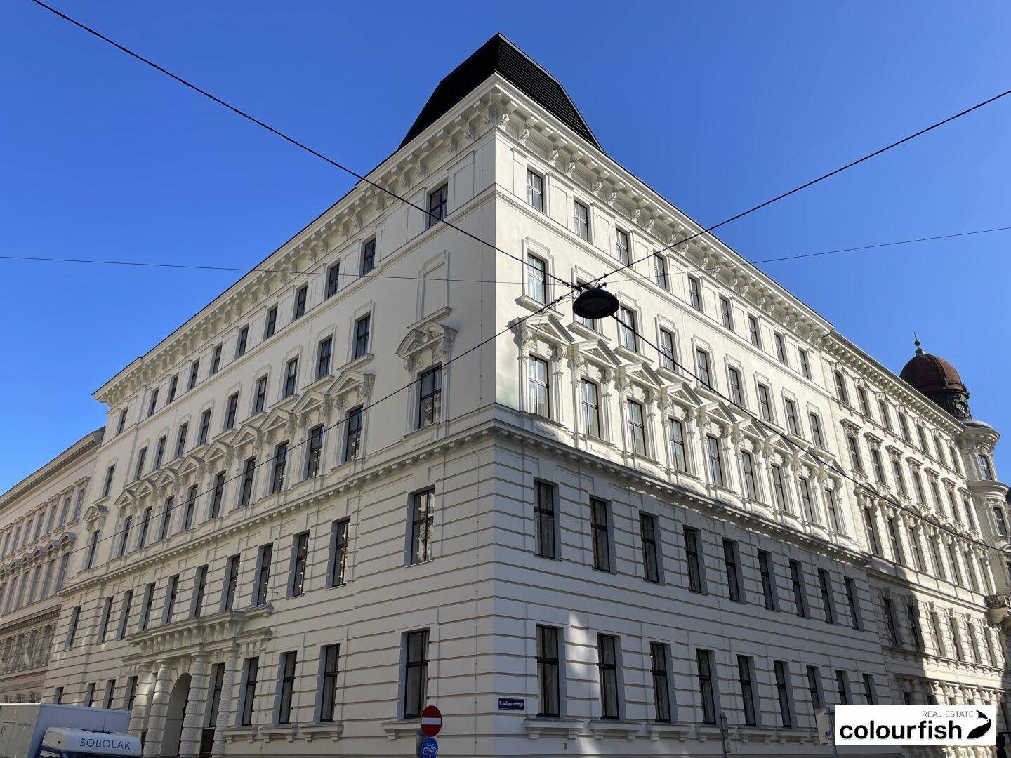 Elegantes Eckgebäude mit klassischer Architektur, vielen Fenstern und einer markanten Dachform unter blauem Himmel.