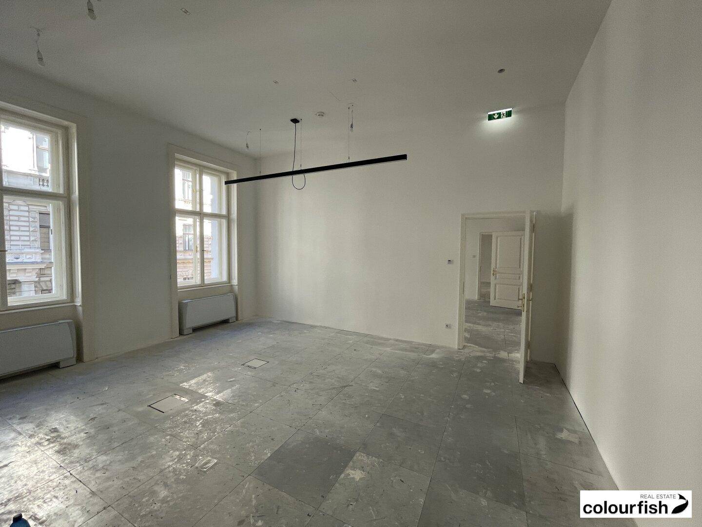 Heller, leerer Raum mit zwei großen Fenstern, unfertigem Boden und moderner Deckenbeleuchtung.