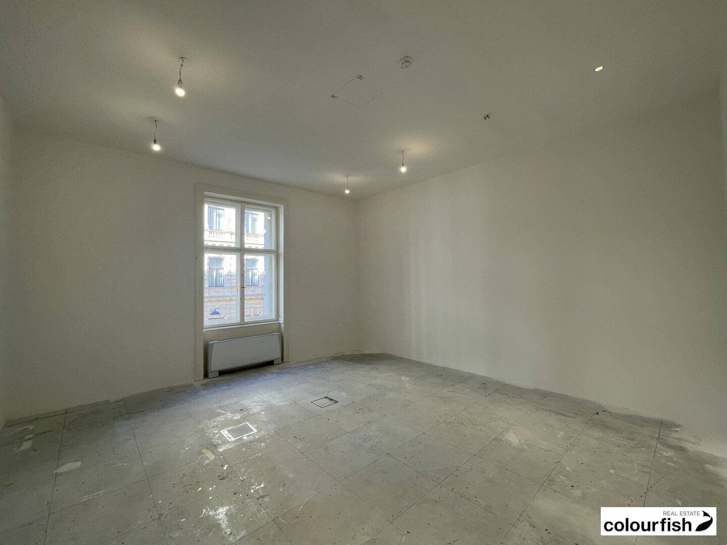 Heller, leerer Raum mit einem großen Fenster, unfertigem Boden und Deckenstrahlern.