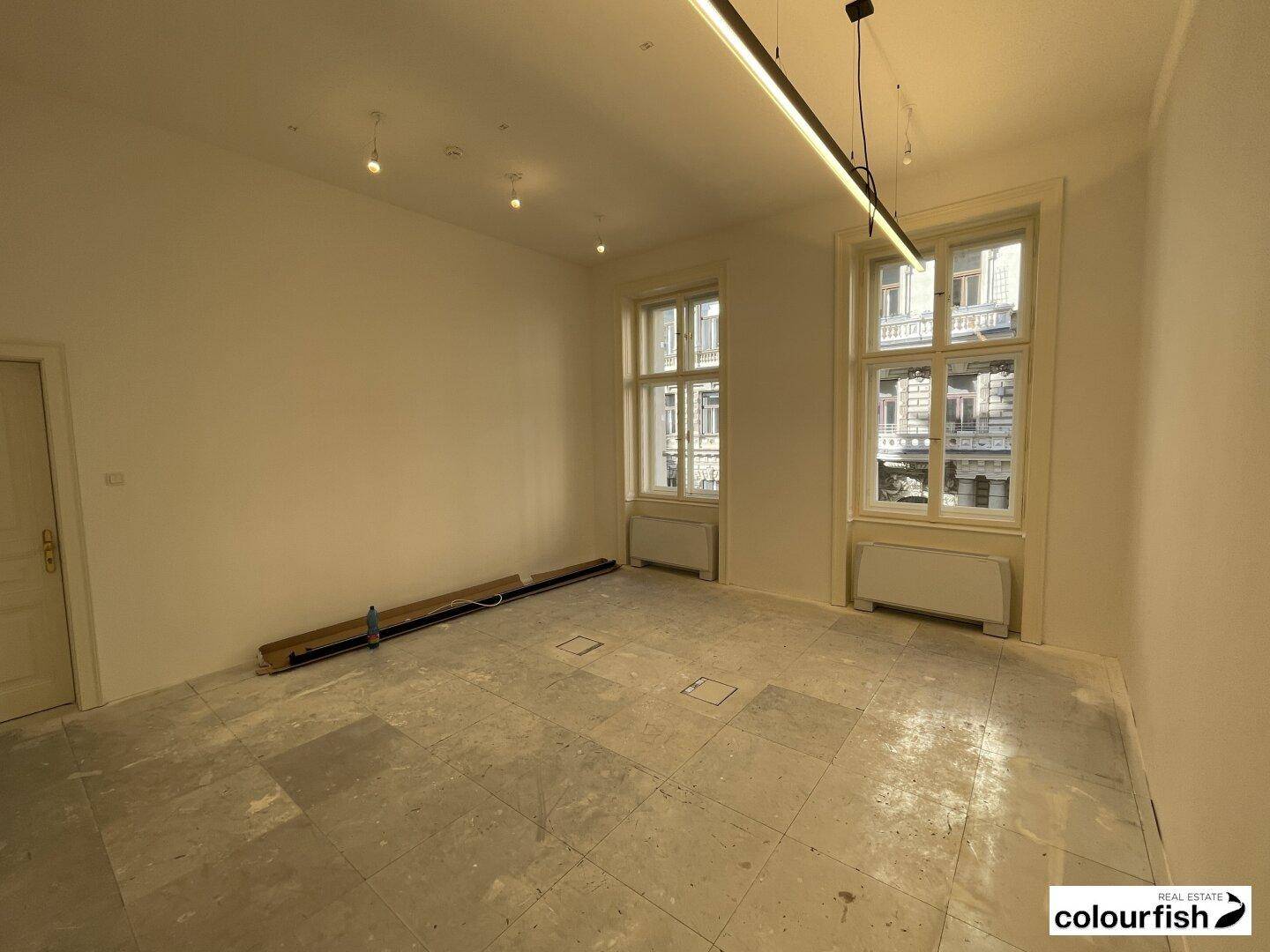 Heller, leerer Raum mit zwei großen Fenstern, unfertigem Boden und moderner Deckenbeleuchtung.