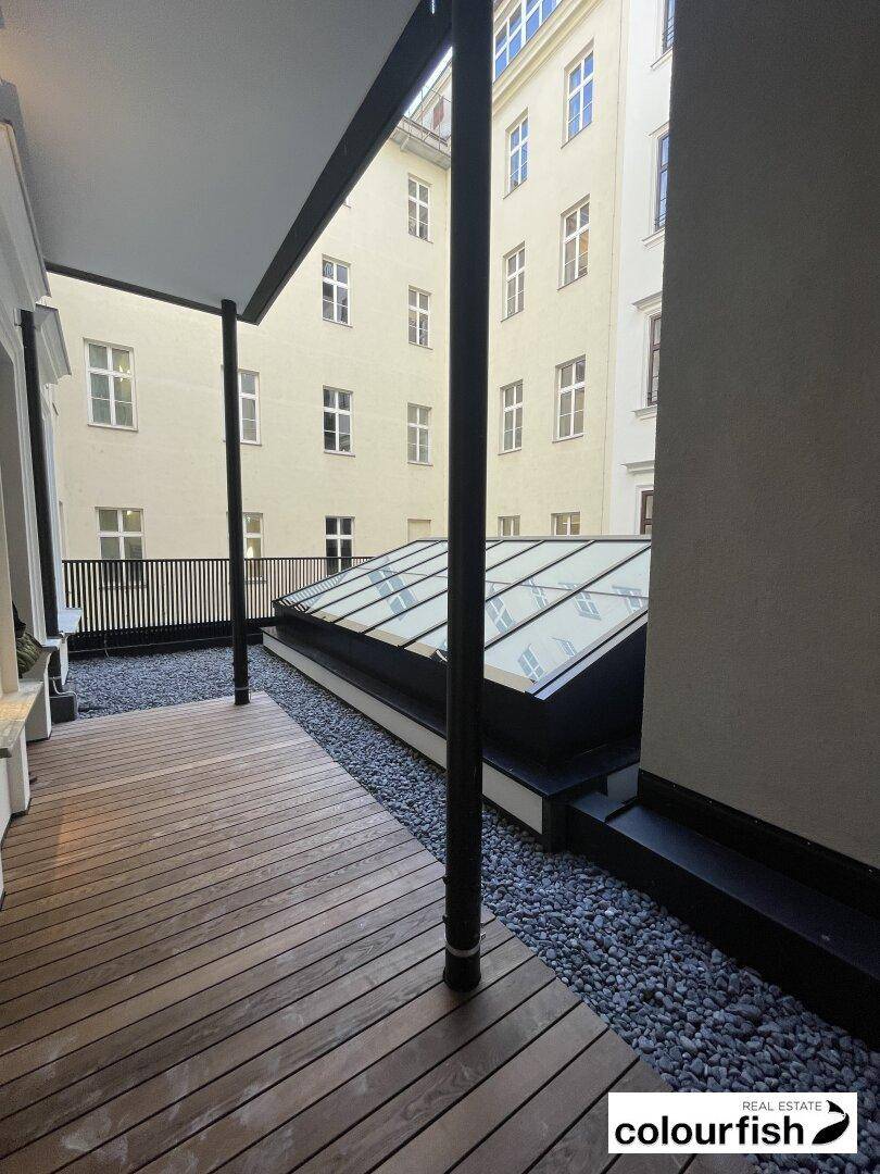 Moderne Terrasse mit Holzdeck, Kiesbereich und Glasdach, umgeben von Gebäudefassaden.