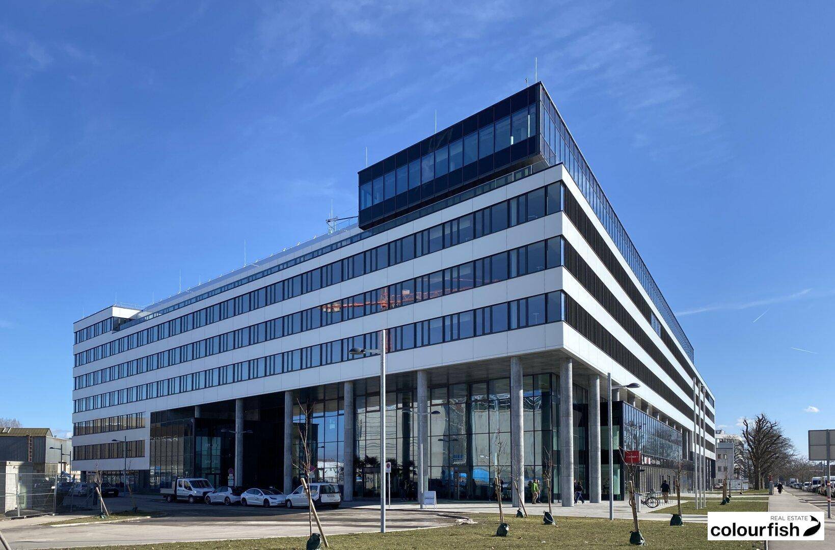 Weitläufiges modernes Bürogebäude mit mehreren Etagen, großen Fenstern und klaren architektonischen Linien unter blauem Himmel.
