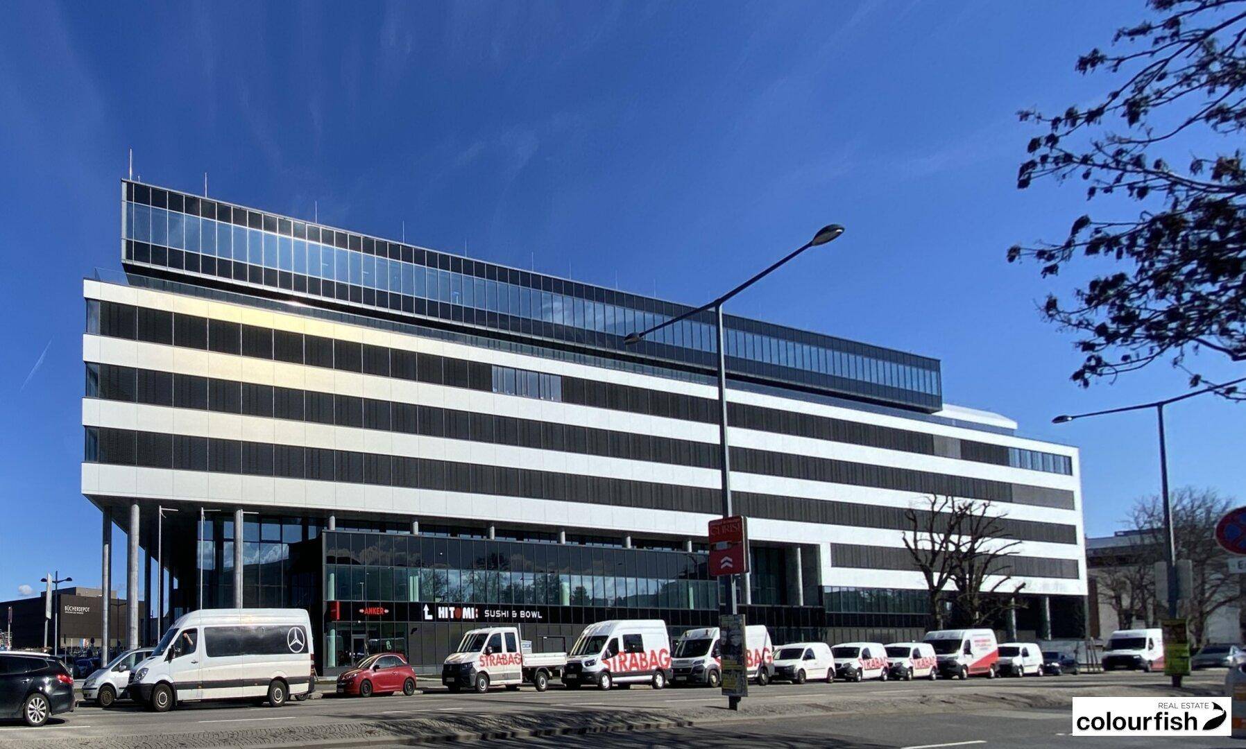 Ansicht eines modernen Bürogebäudes von der Straße aus, mit parkenden Fahrzeugen und Geschäften im Erdgeschoss.