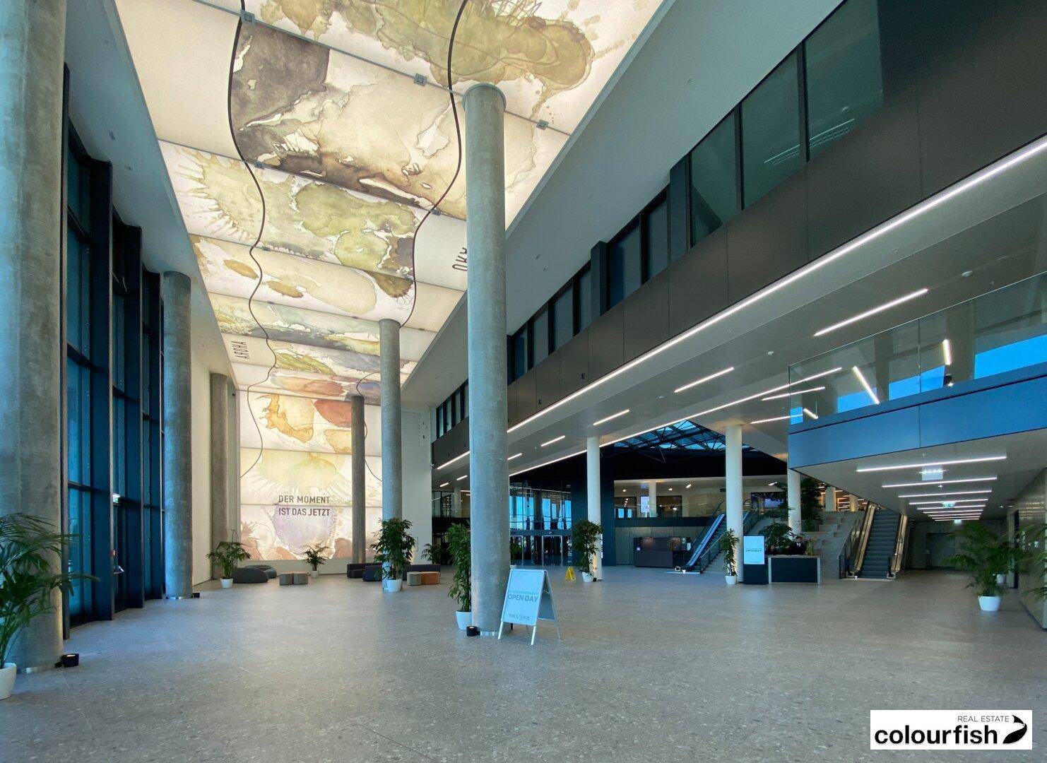 Geräumige, moderne Lobby mit hohen Decken, großen Säulen und einer kunstvollen Deckenbeleuchtung, die eine offene Atmosphäre schafft.