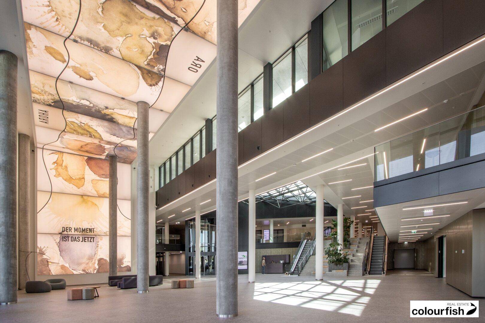 Geräumige, moderne Lobby mit hohen Decken, künstlerischer Deckengestaltung und viel Tageslicht.