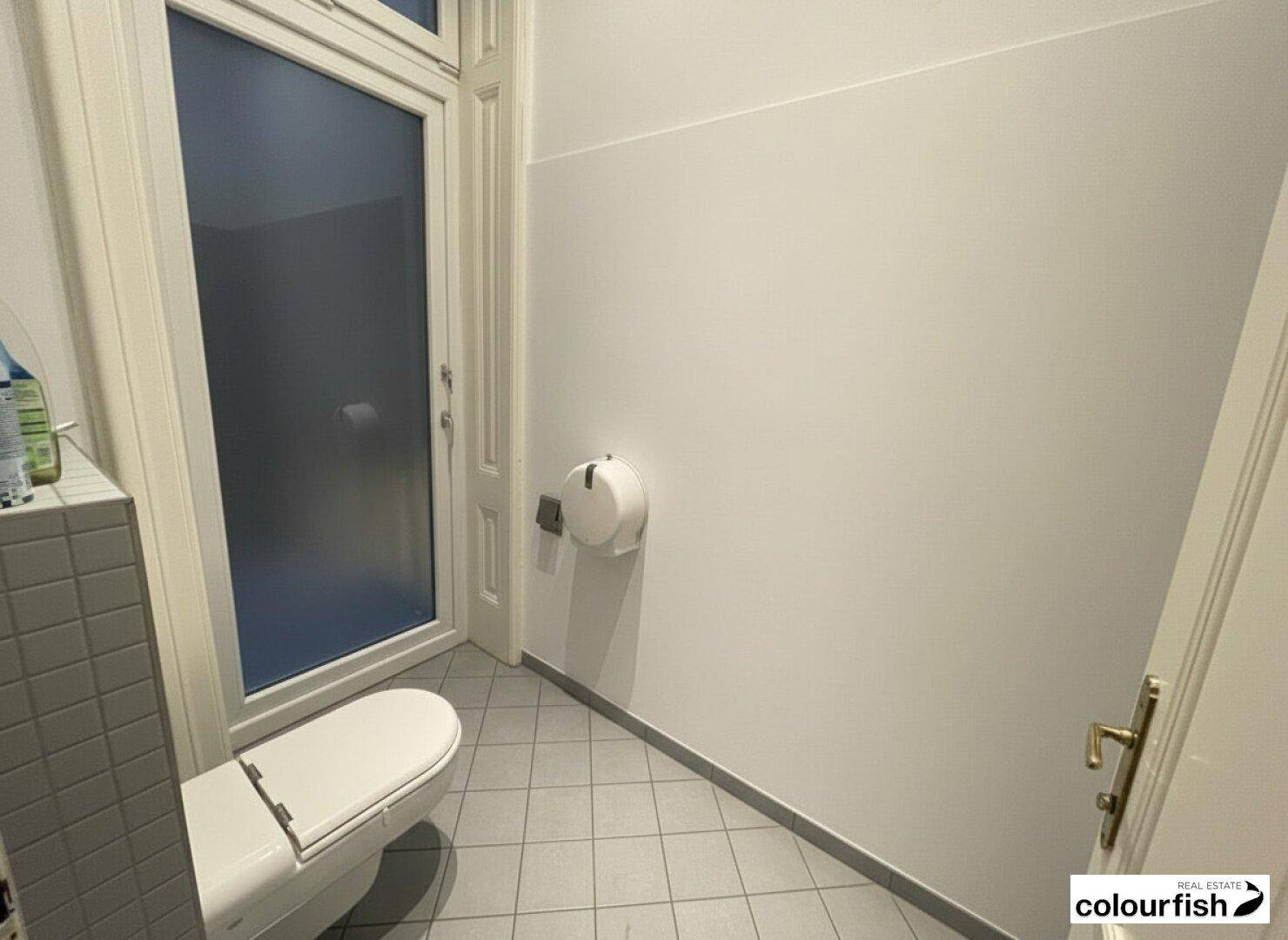 Separate Toilette mit grauen Bodenfliesen, wandhängendem WC und einem Milchglasfenster.