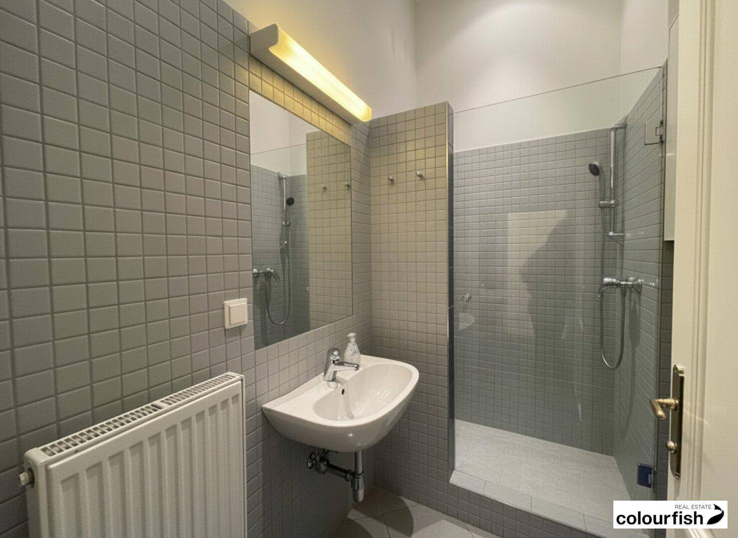 Modernes Badezimmer mit grauen Wandfliesen, Duschkabine und einem Waschbecken mit Spiegel.