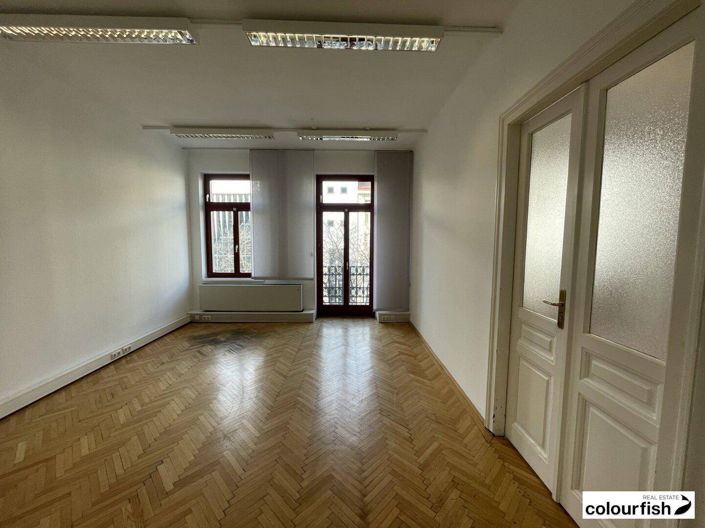Geräumiger Büroraum mit glänzendem Fischgrätparkett, weißen Wänden und Zugang zu einem kleinen Balkon.