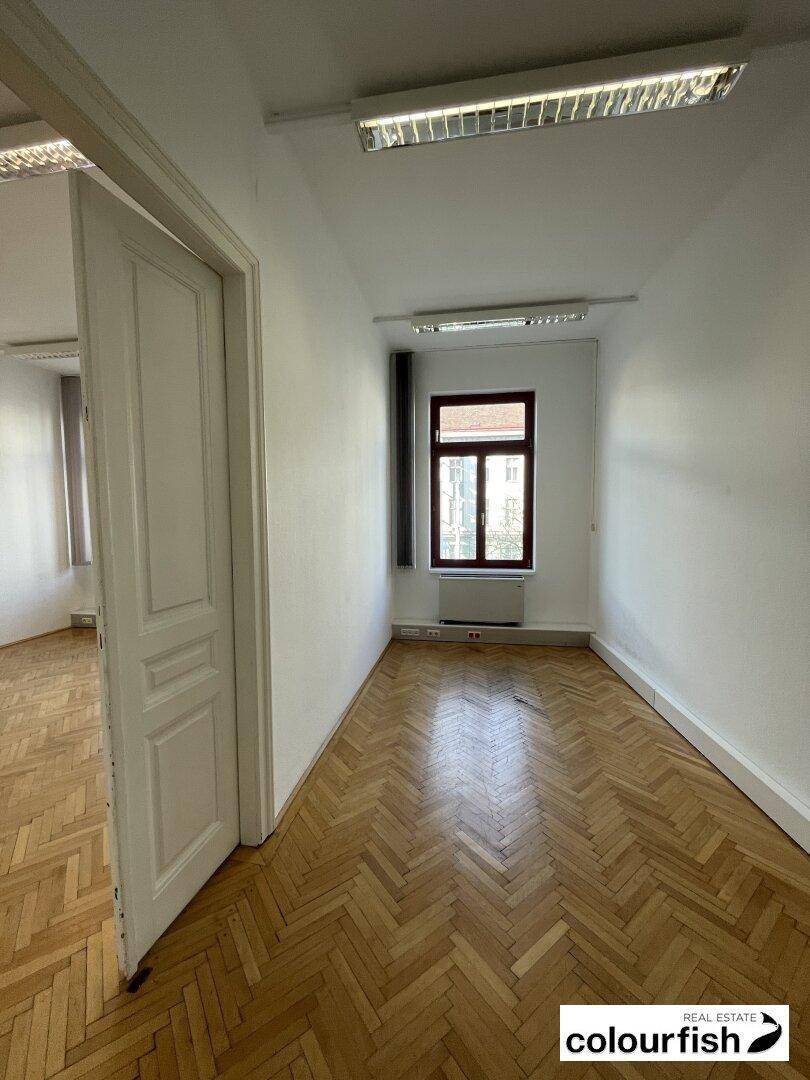 Schmaler Büroraum mit glänzendem Fischgrätparkett, einem Fenster und weißen Wänden, ideal für ein Einzelbüro.