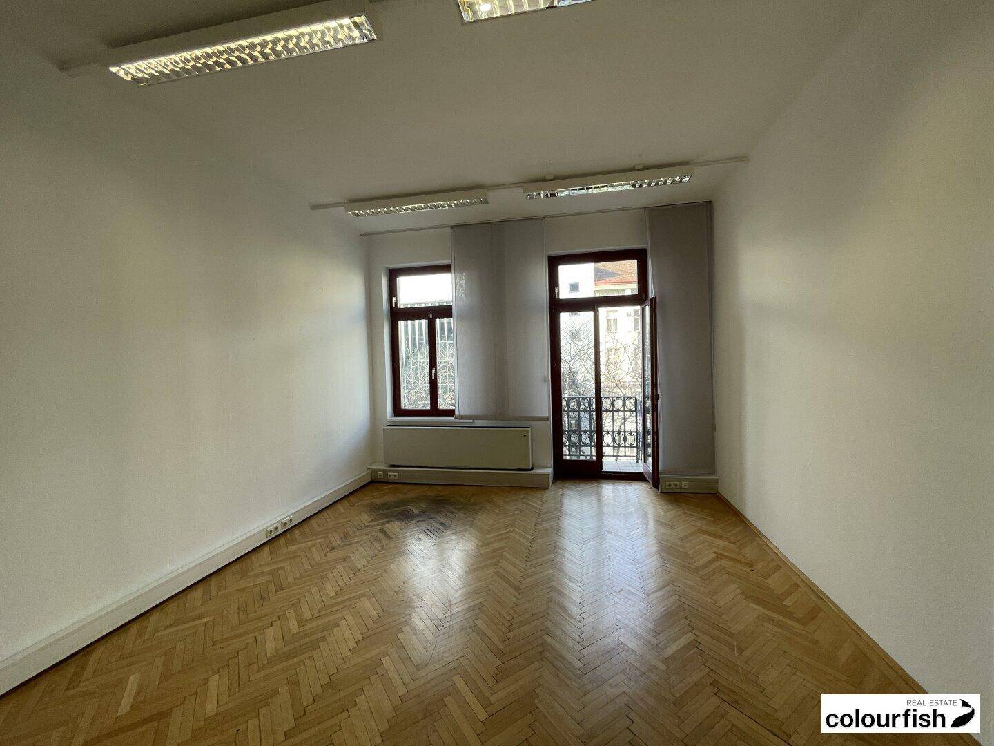 Geräumiger Büroraum mit glänzendem Fischgrätparkett, weißen Wänden und Zugang zu einem kleinen Balkon.