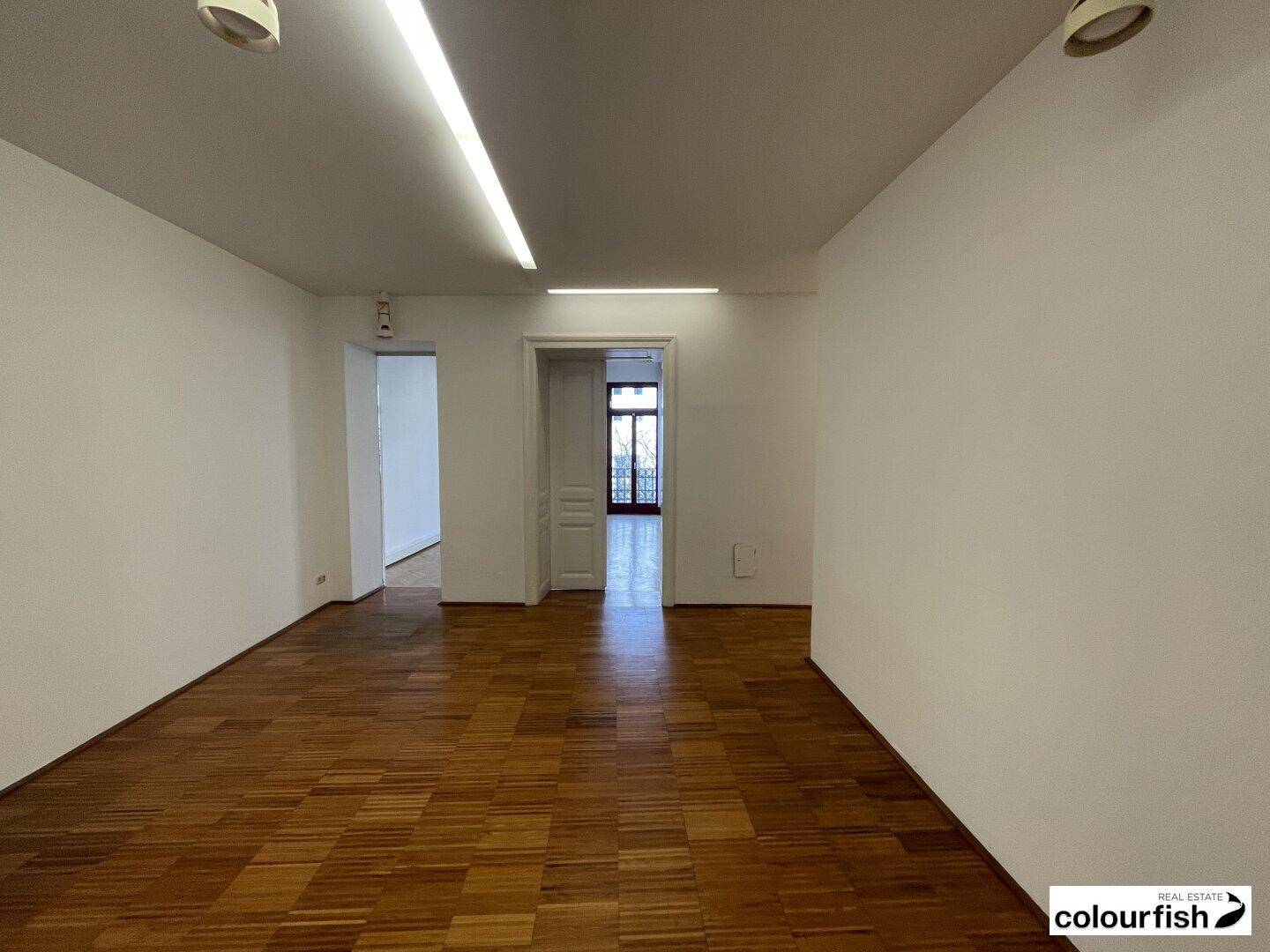 Langer Büroraum mit glänzendem Holzparkett, weißen Wänden und Blick auf einen weiteren Raum.