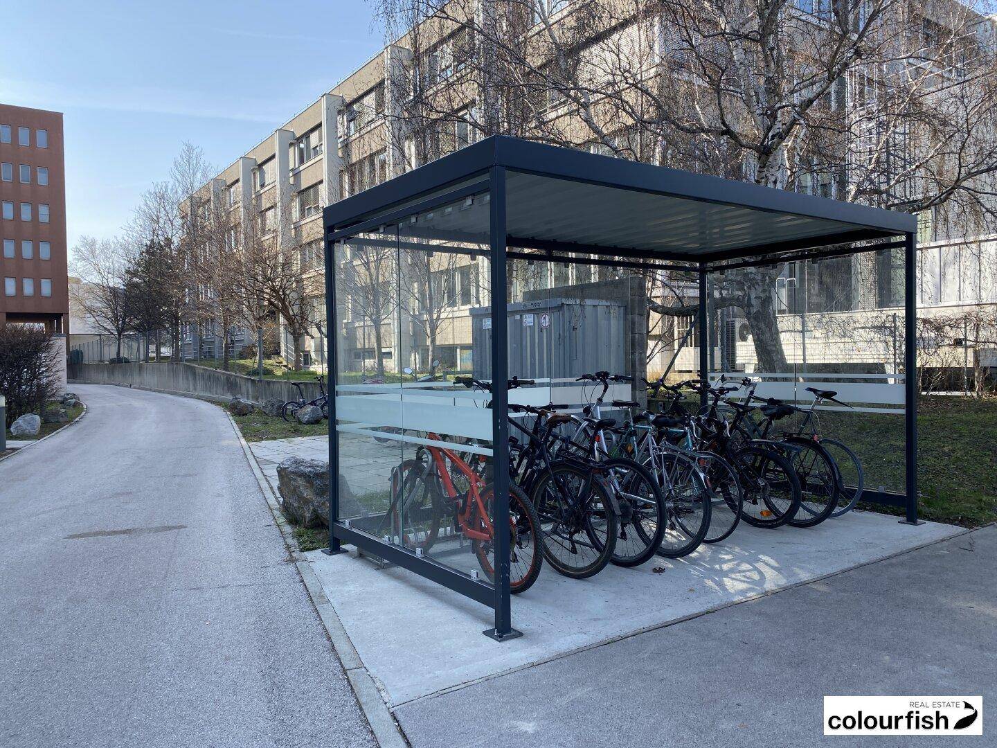 Überdachter Fahrradunterstand mit mehreren Fahrrädern vor einem Bürogebäude.