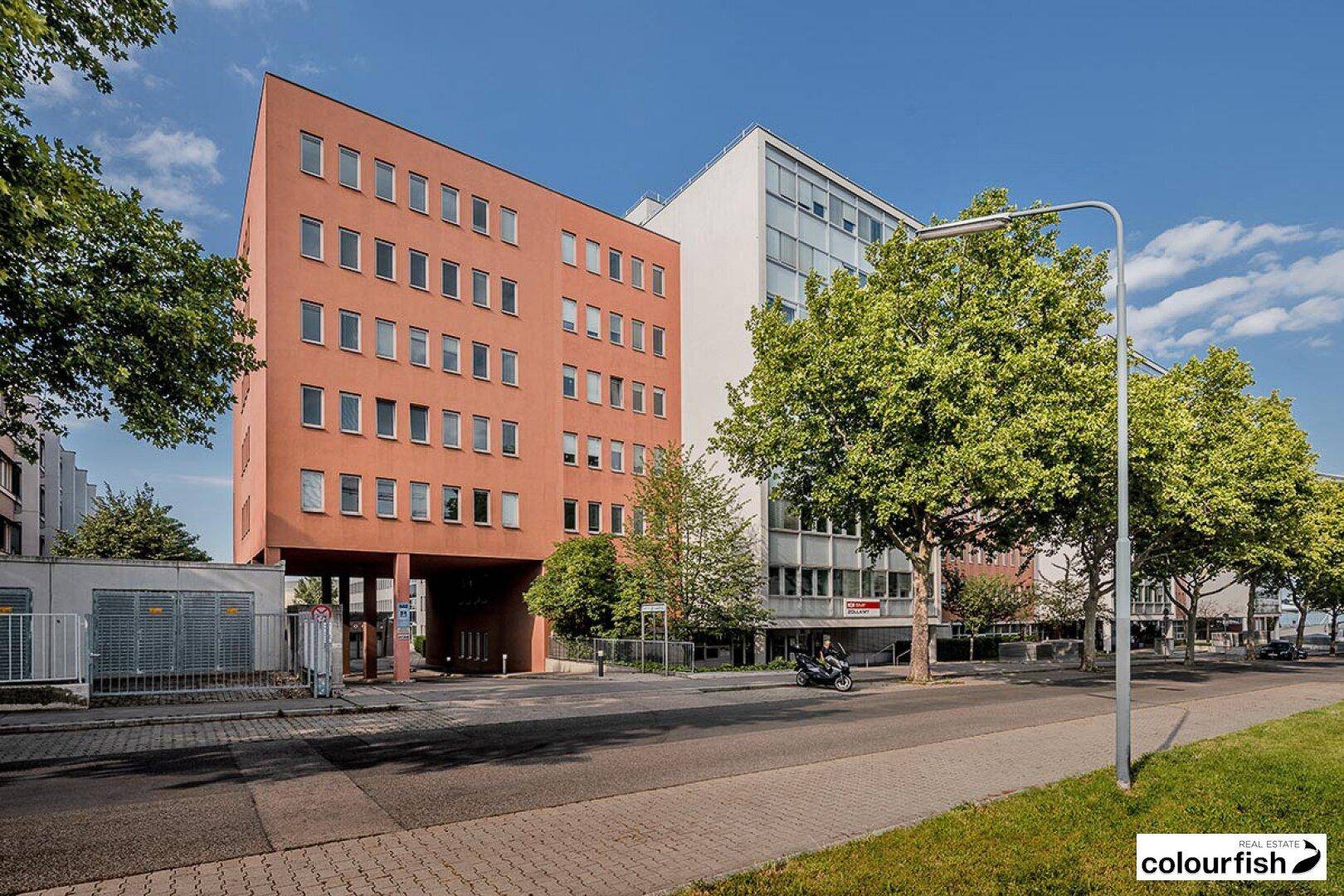Farbige Gebäudefassade in Rot-Orange neben einem hellen Gebäude an einer städtischen Straße mit Bäumen.
