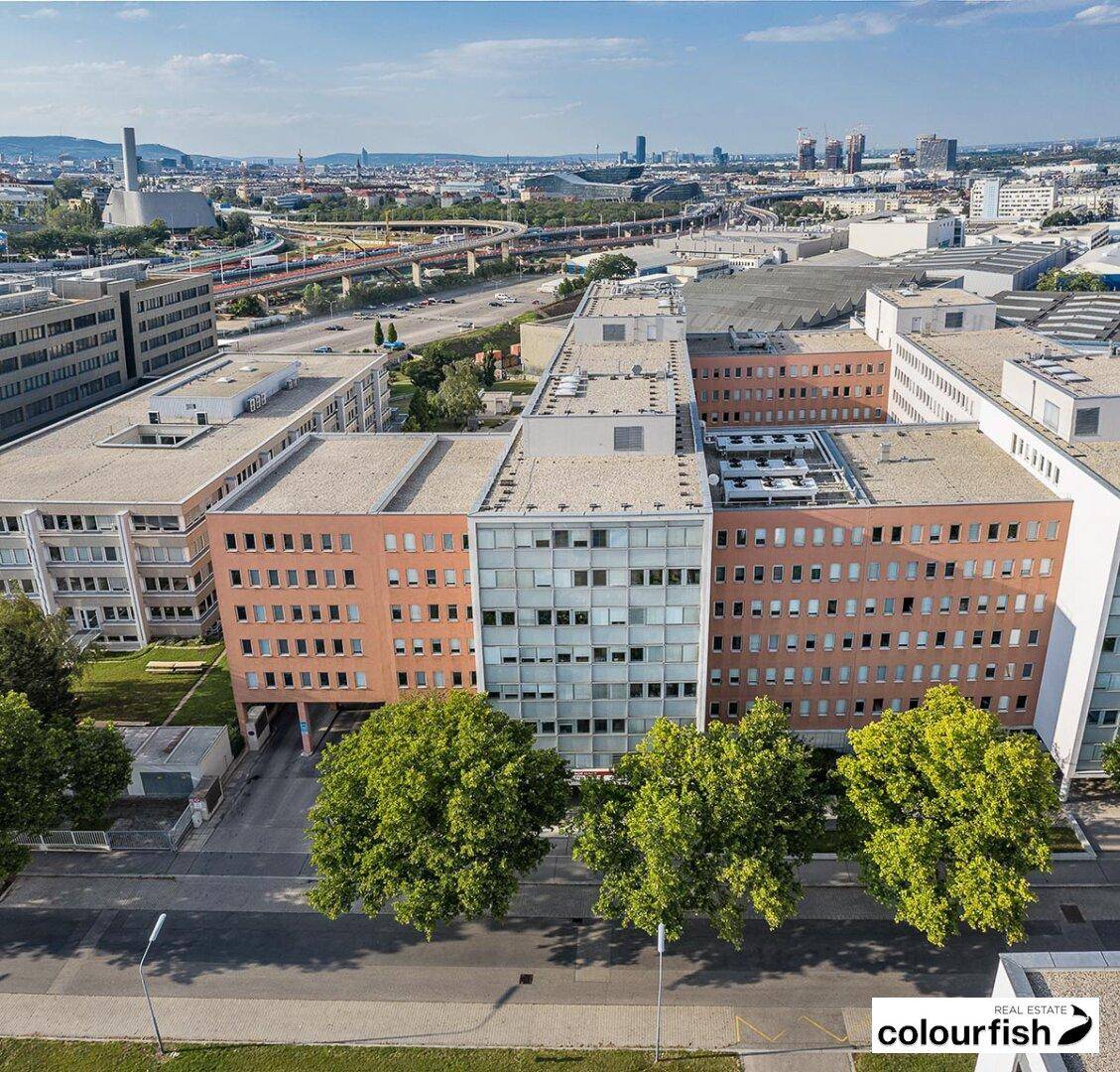 Luftaufnahme eines modernen Gebäudekomplexes mit verschiedenen Fassadenfarben und städtischer Infrastruktur im Hintergrund.