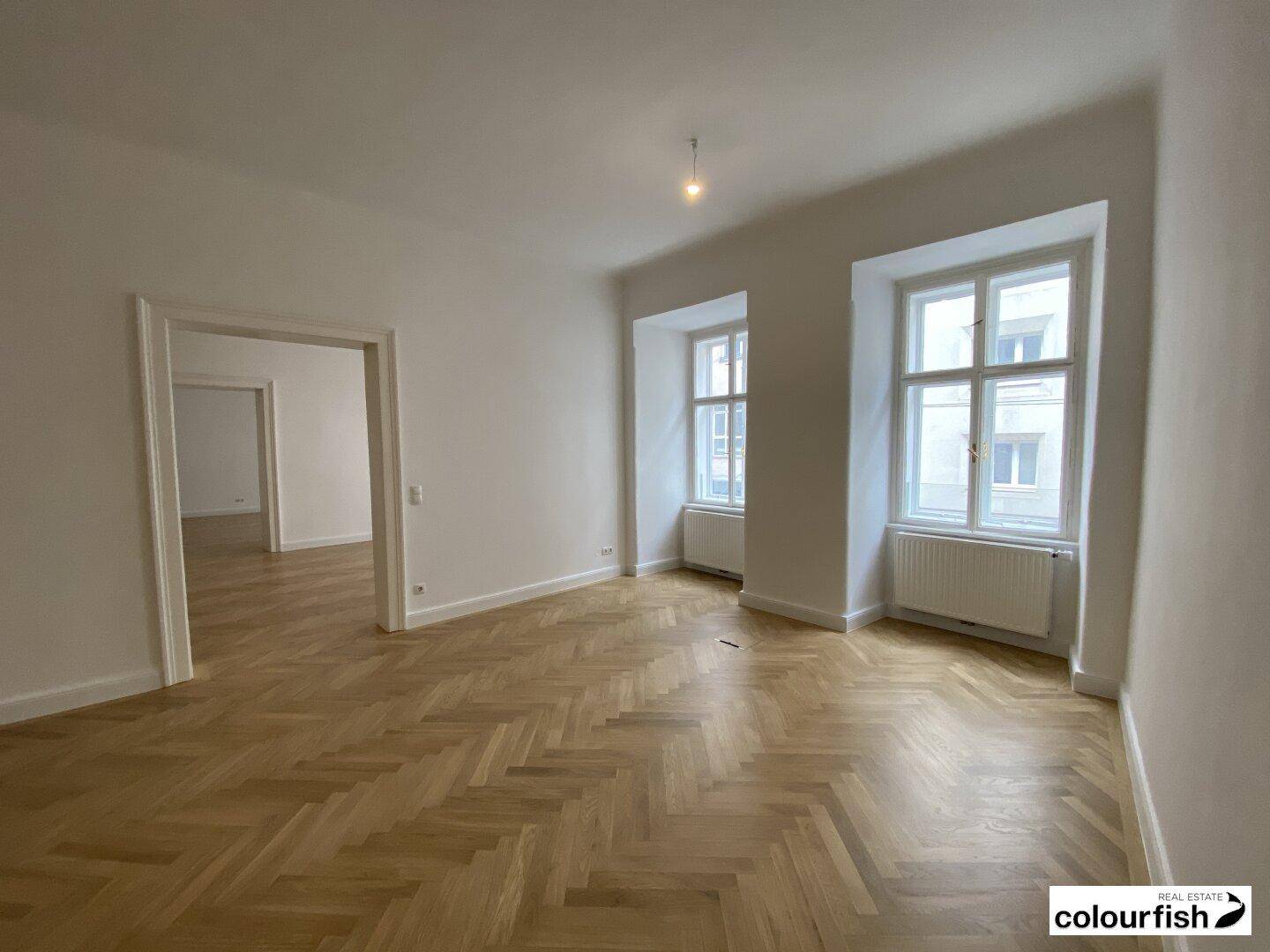 Großer, heller Raum mit Fischgrätparkett und hohen Decken, durch Doppeltüren mit weiteren Räumen verbunden.